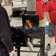 Portable metal fire pit with orange flames and charcoal, surrounded by people in red shirts outdoors.