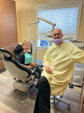 A dentist in yellow gown and a young patient smile in a dental office.