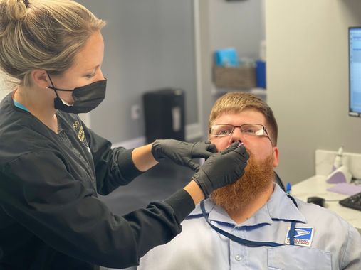 Dental assistant, in black mask and gloves, examines a man's mouth in an office setting.