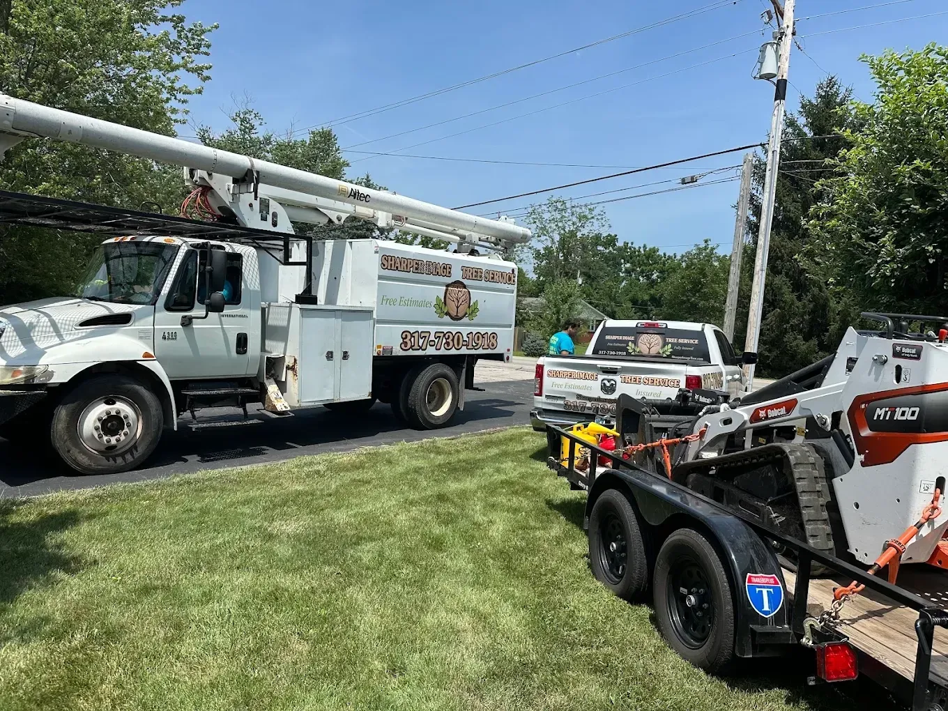 Utility trucks and equipment trailers parked on a grassy roadside near overhead power lines under a blue sky