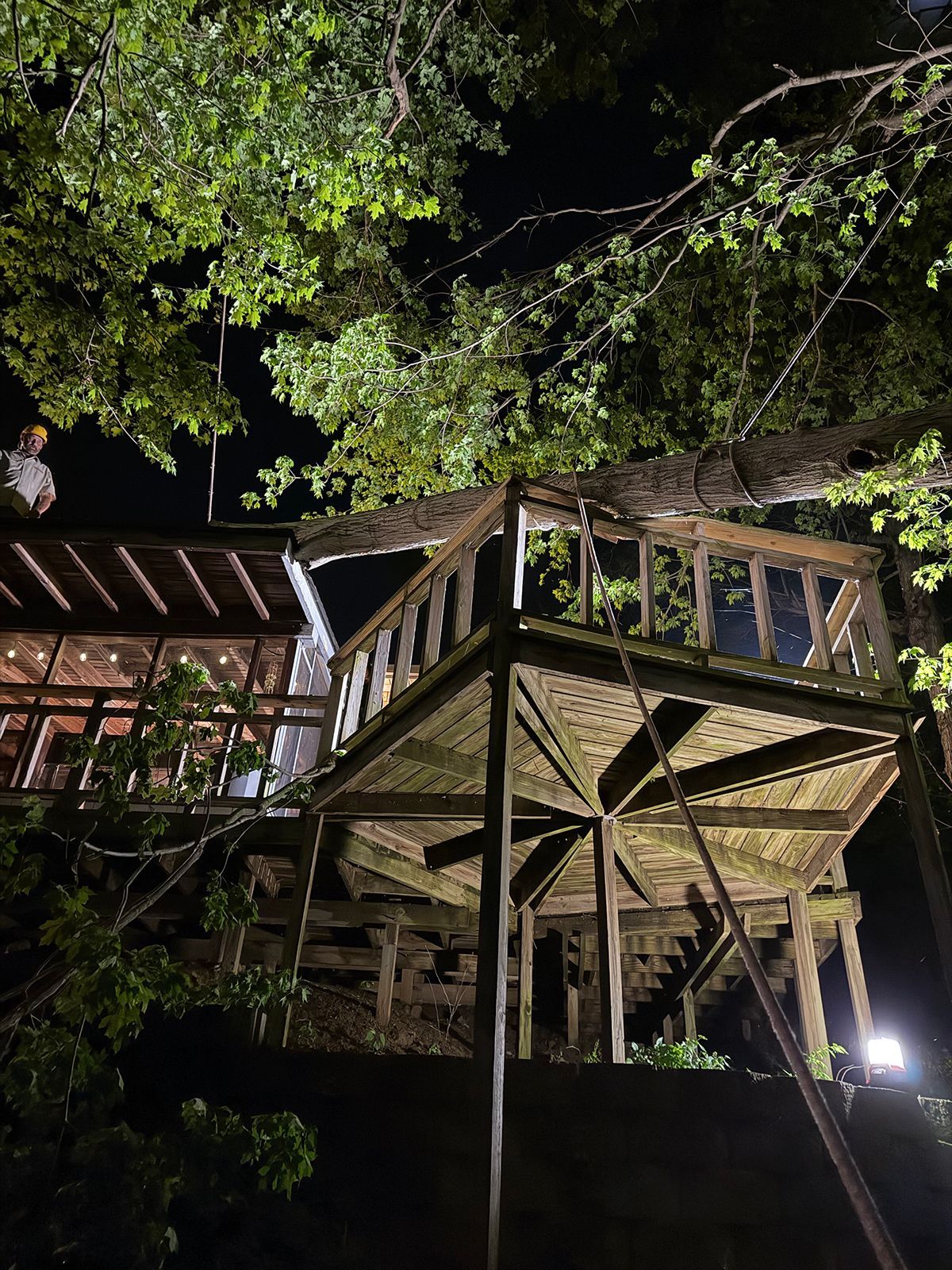 Nighttime view of a wooden treehouse deck with stairs, lit from below and surrounded by leafy branches