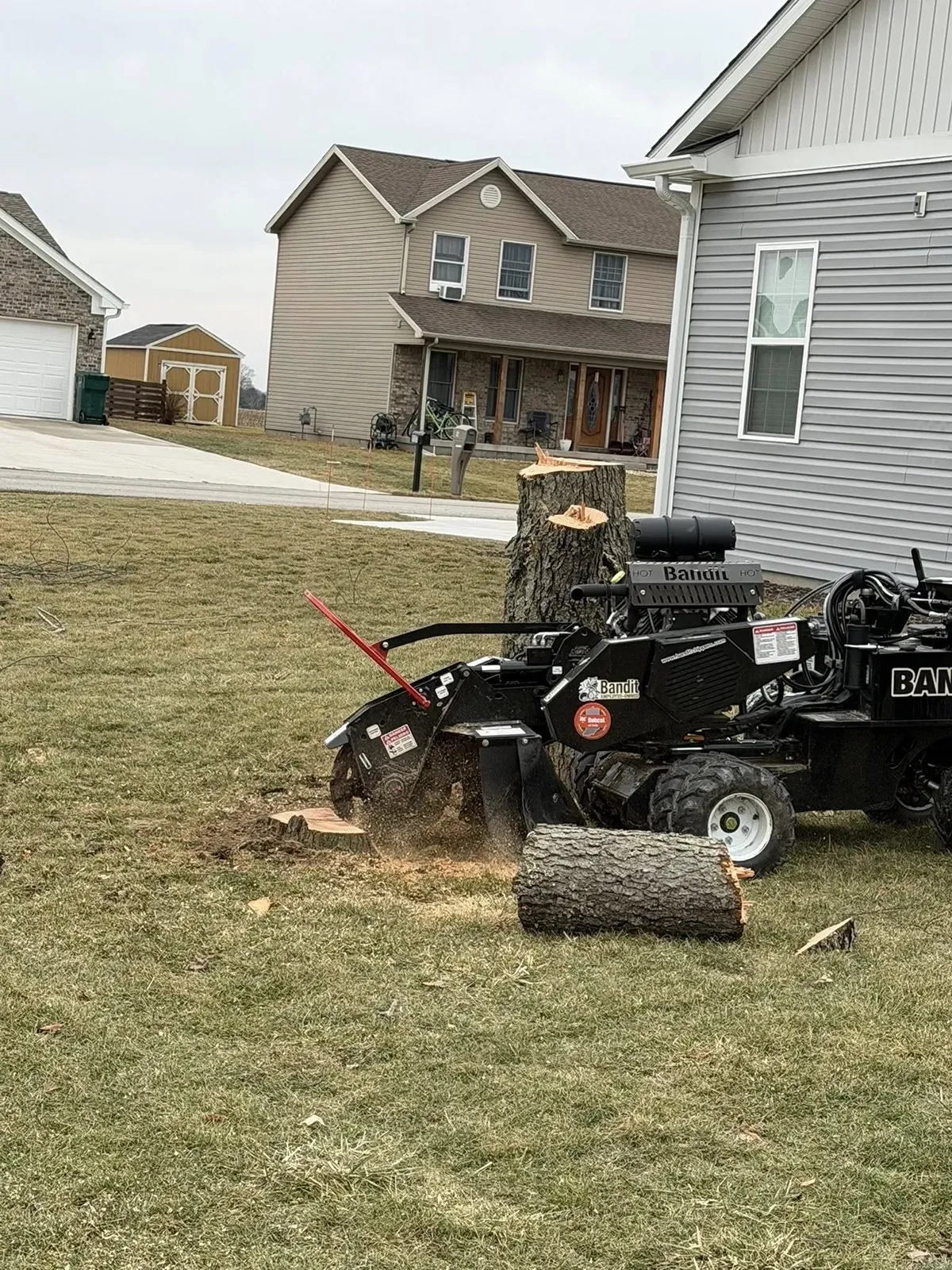Lawn mower clearing grass near a house, with a cut tree stump and wood chip debris in the yard.
