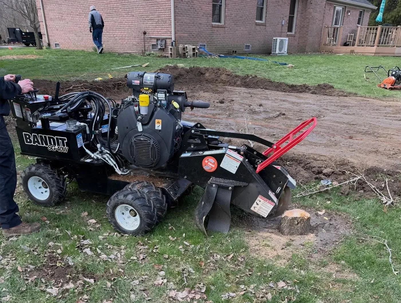 Black tow-behind stump grinder attached to a machine in a muddy yard near a brick house.
