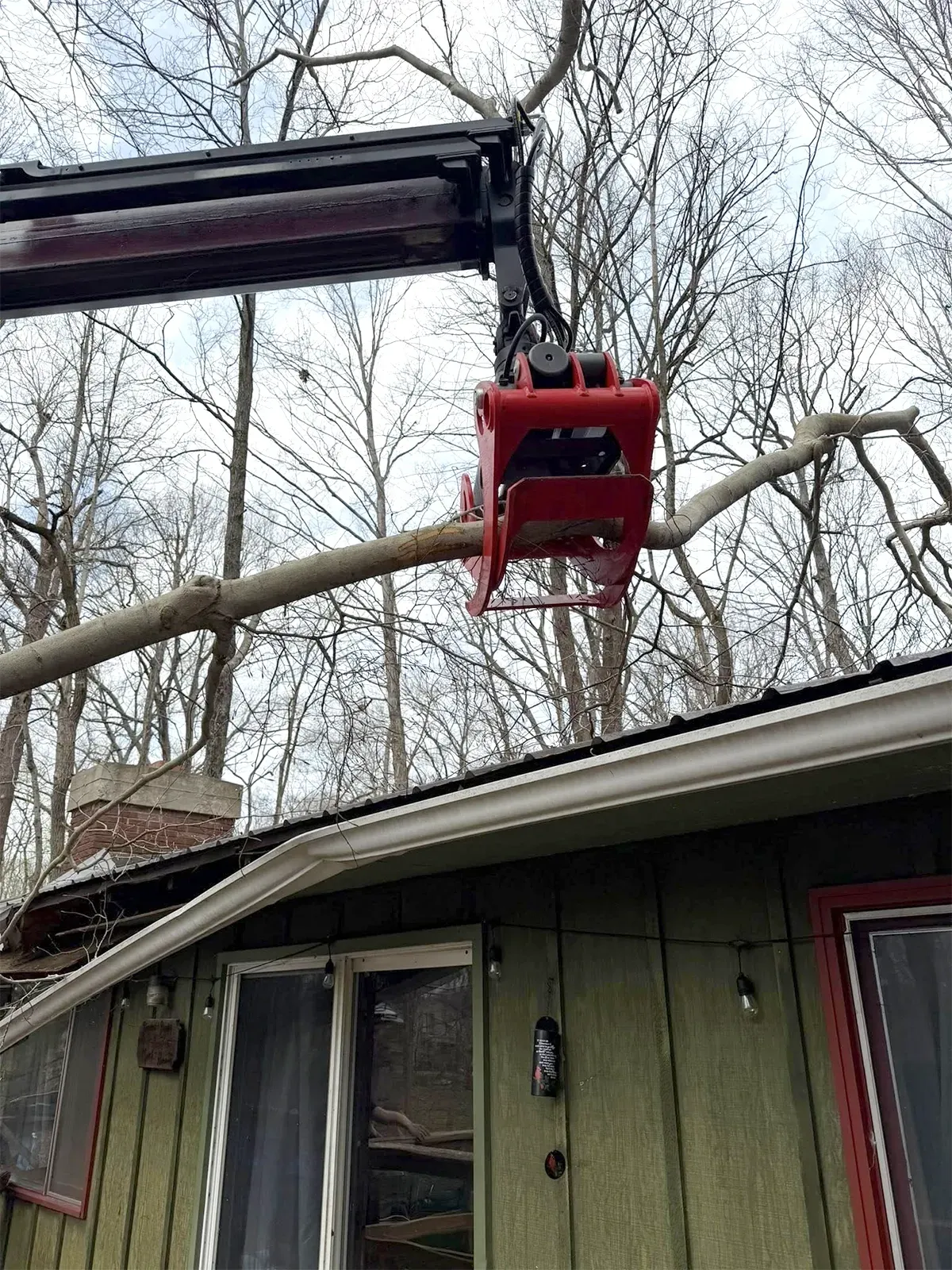 Red crane lifting a fallen tree limb over a house roof in a wooded yard