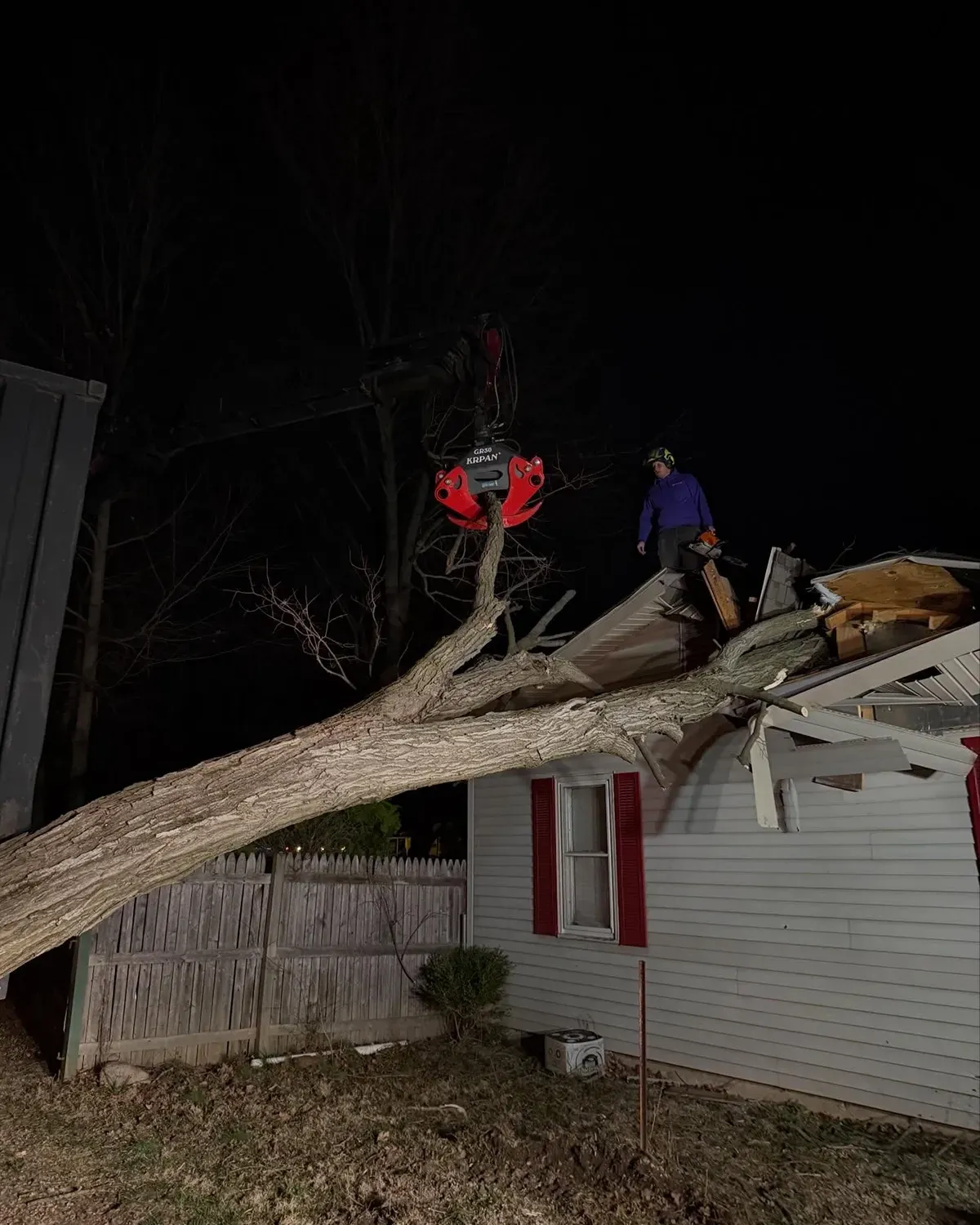 Two workers stand on a fallen tree across a house roof at night after storm damage.