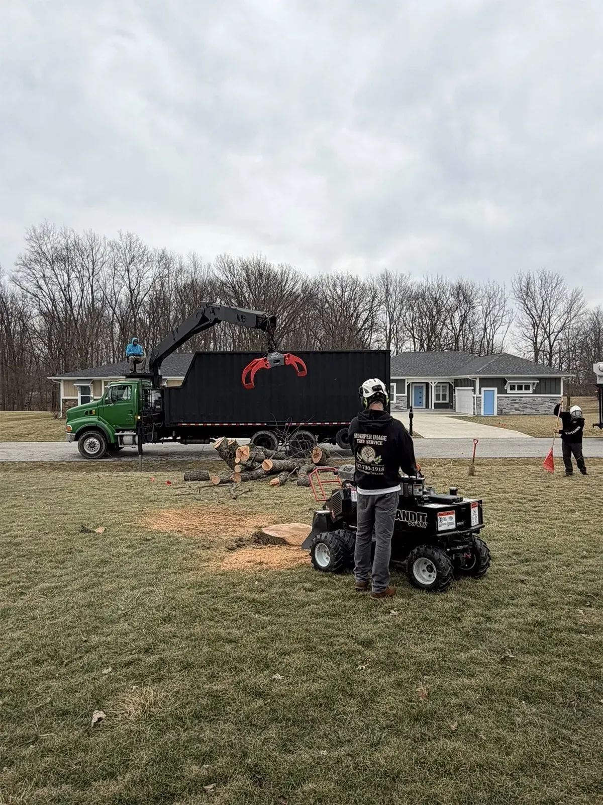 Workers operating a crane truck and ATVs on a grassy field near houses