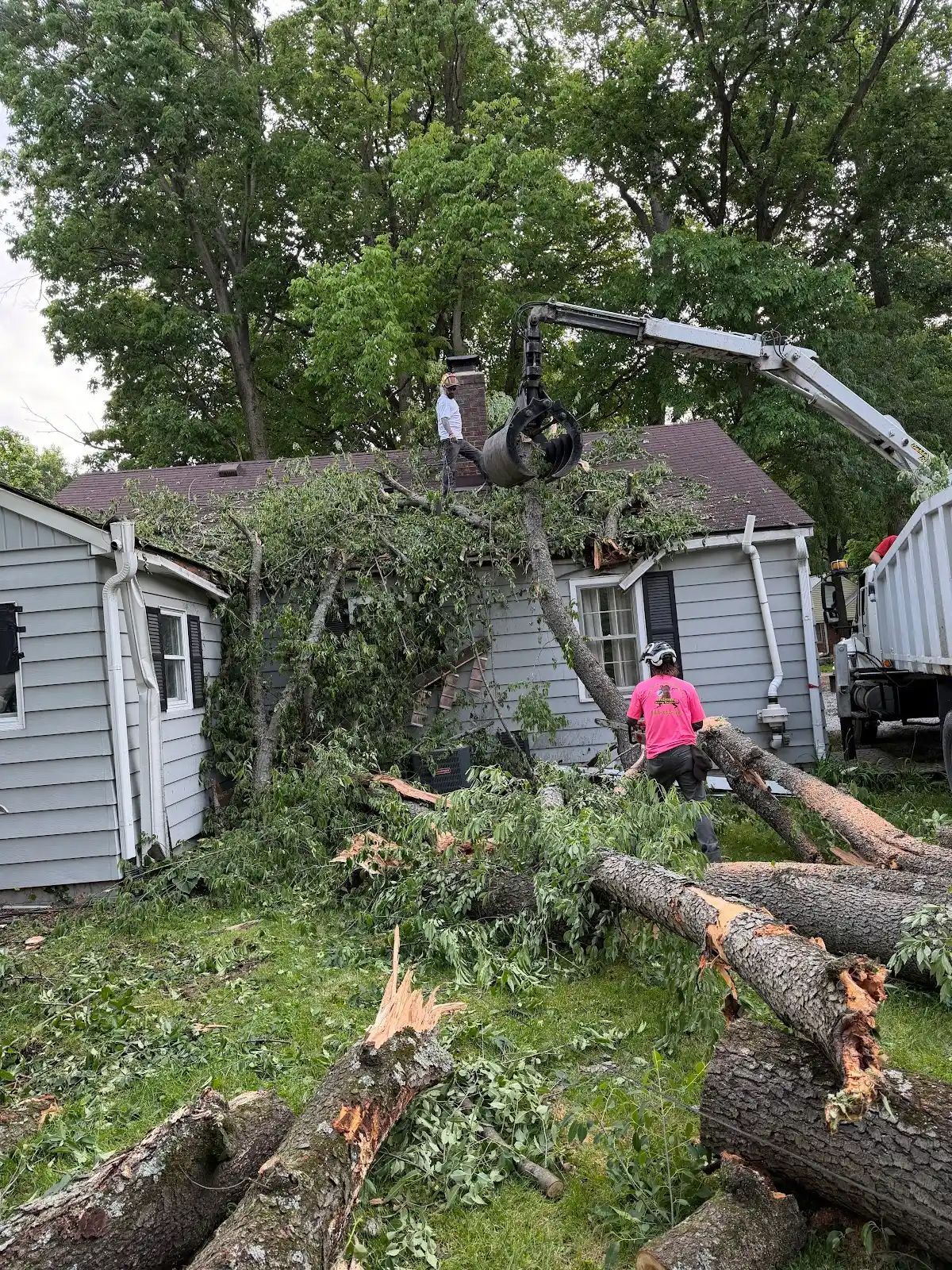 Storm-damaged yard with fallen tree limbs, a house, and a worker using a bucket truck to clear debris
