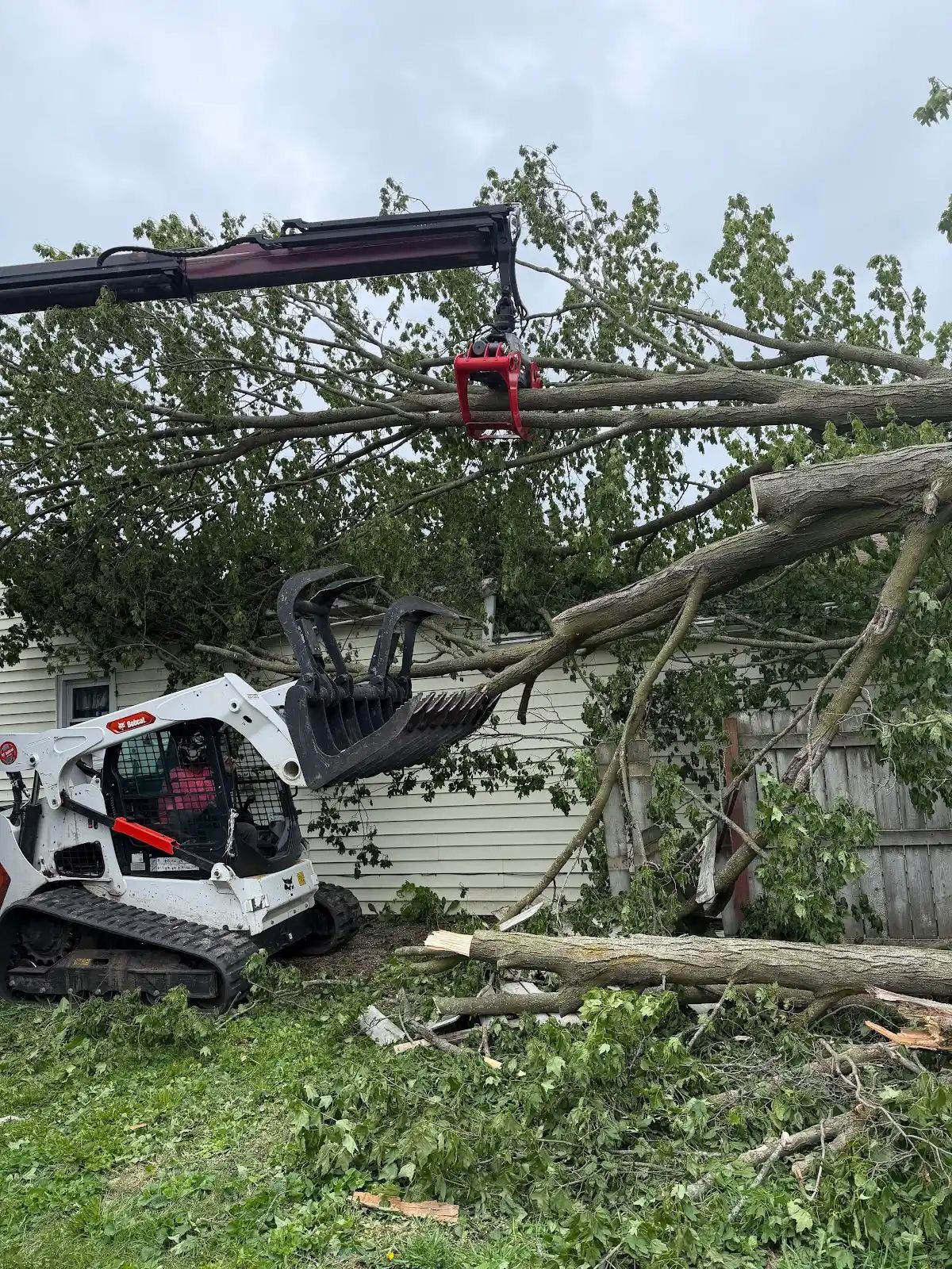 Bobcat loader clearing fallen trees beside a house after storm damage, with a worker on the boom