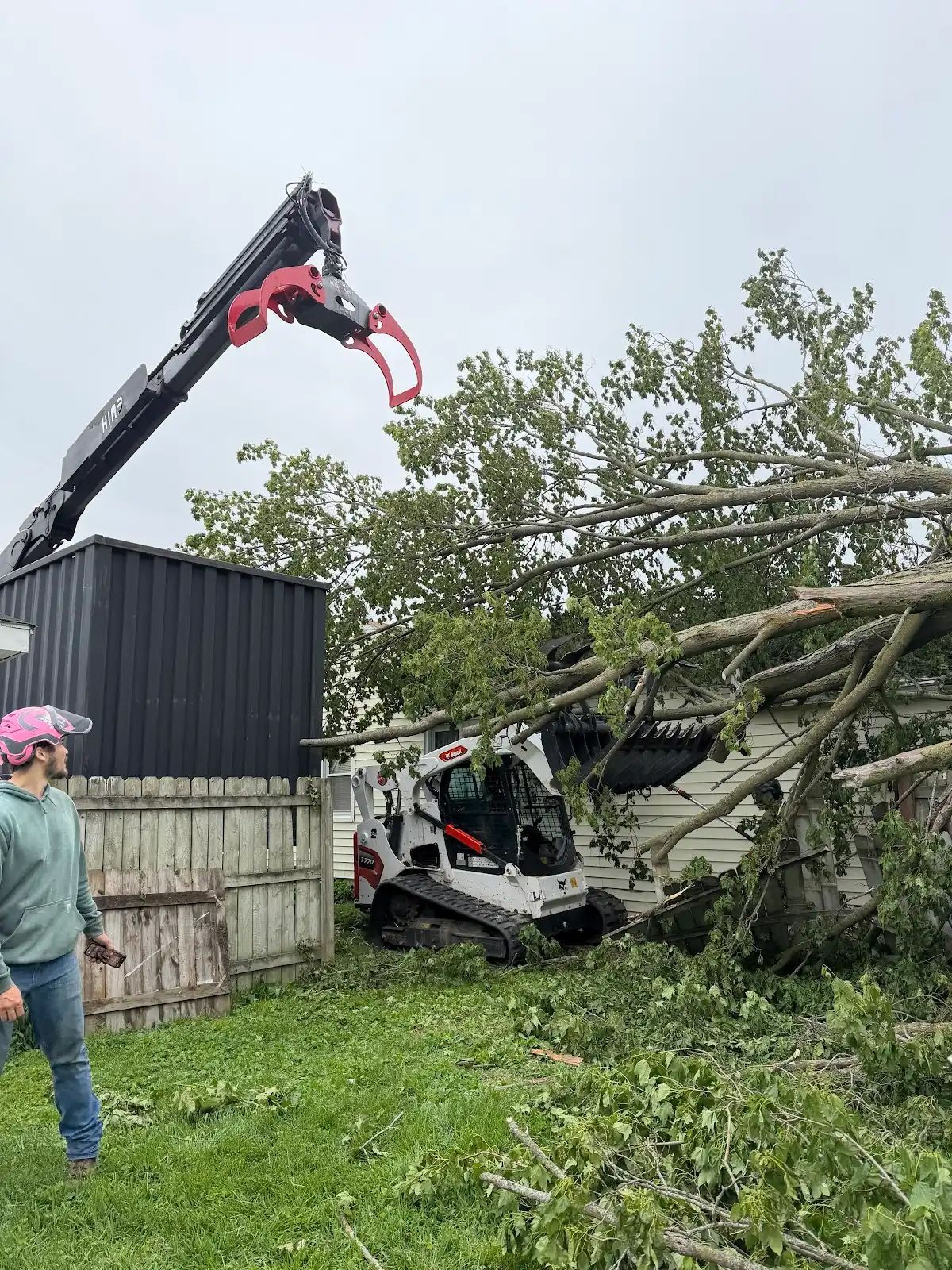 Tree-trimming crane lifting a fallen tree off a white utility vehicle in a grassy yard