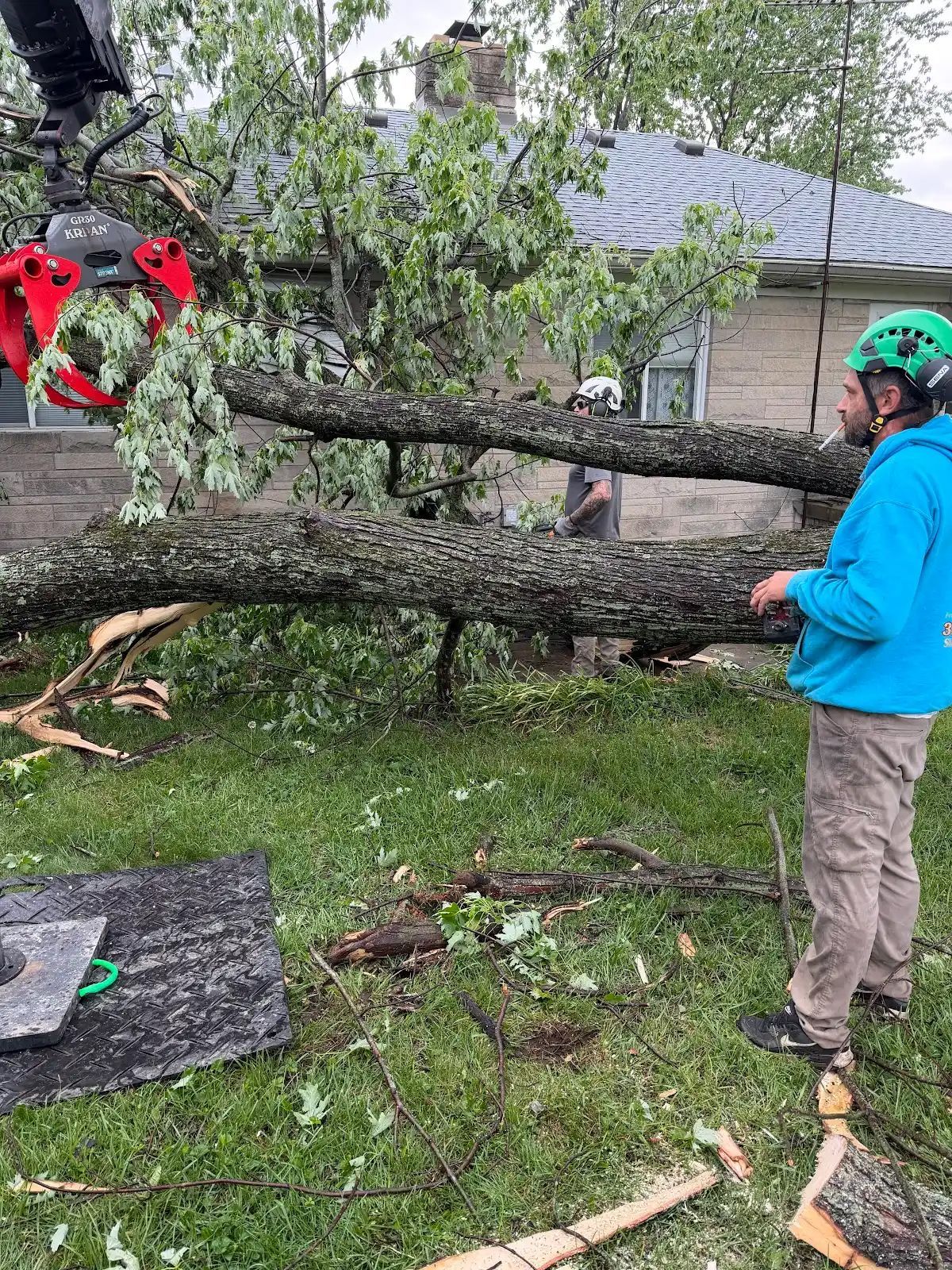 Worker in blue jacket and green helmet cutting a fallen tree limb in a yard