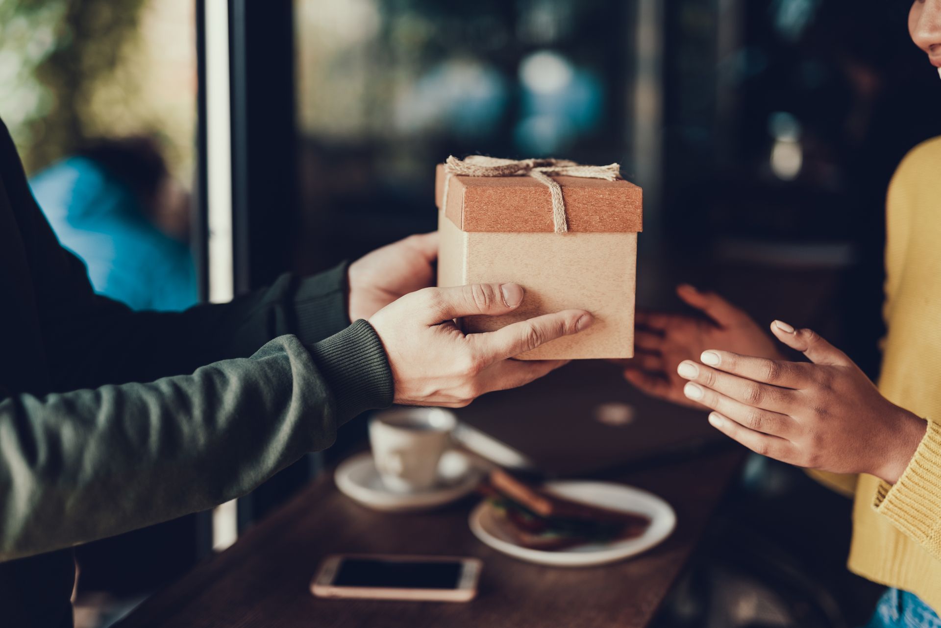 Hands exchanging a wrapped gift box across a table in a cozy indoor setting