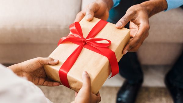 Hands passing a wrapped gift box with a red ribbon on a couch.