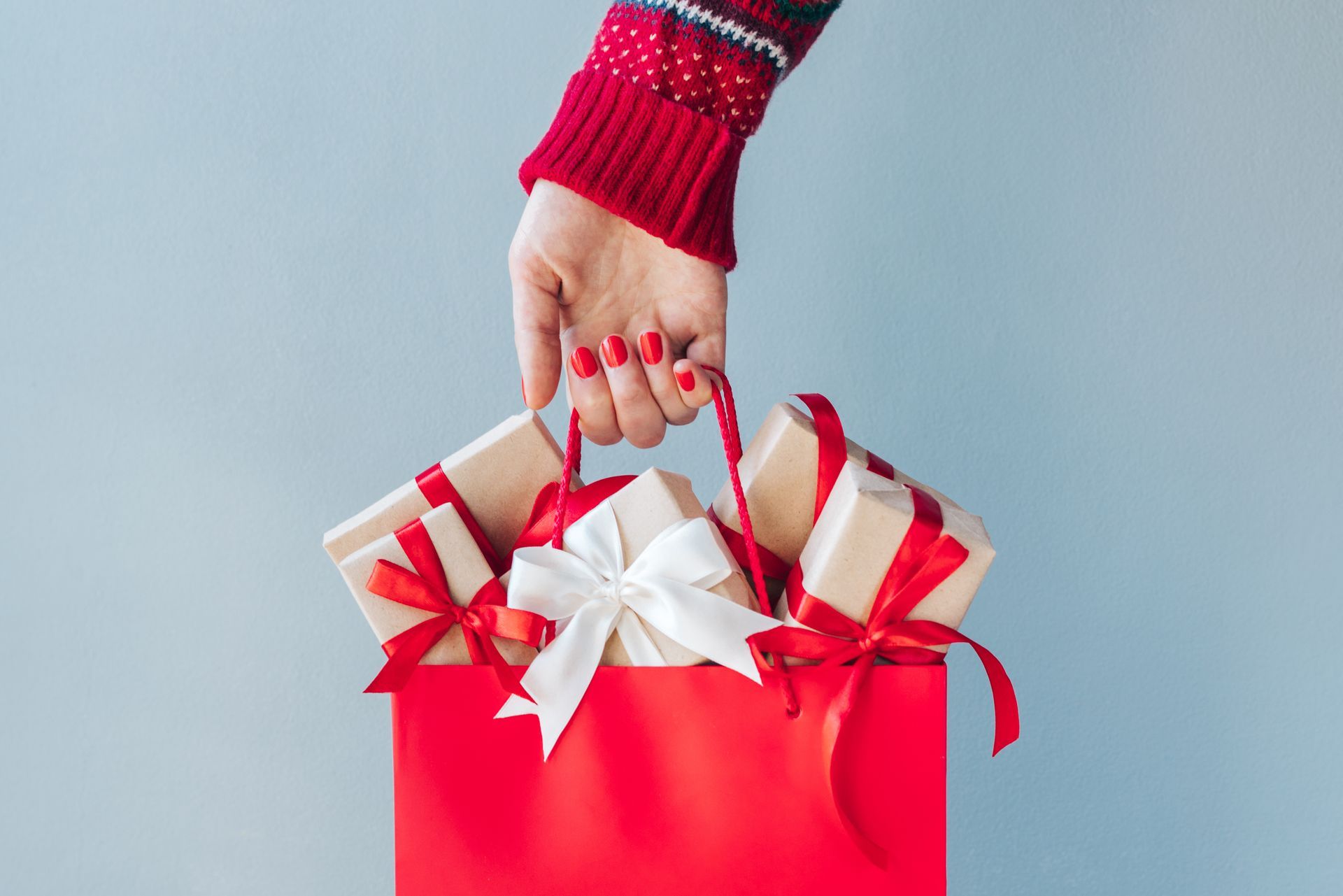 Hand holding red gift bag with white ribbon bows on a light blue background