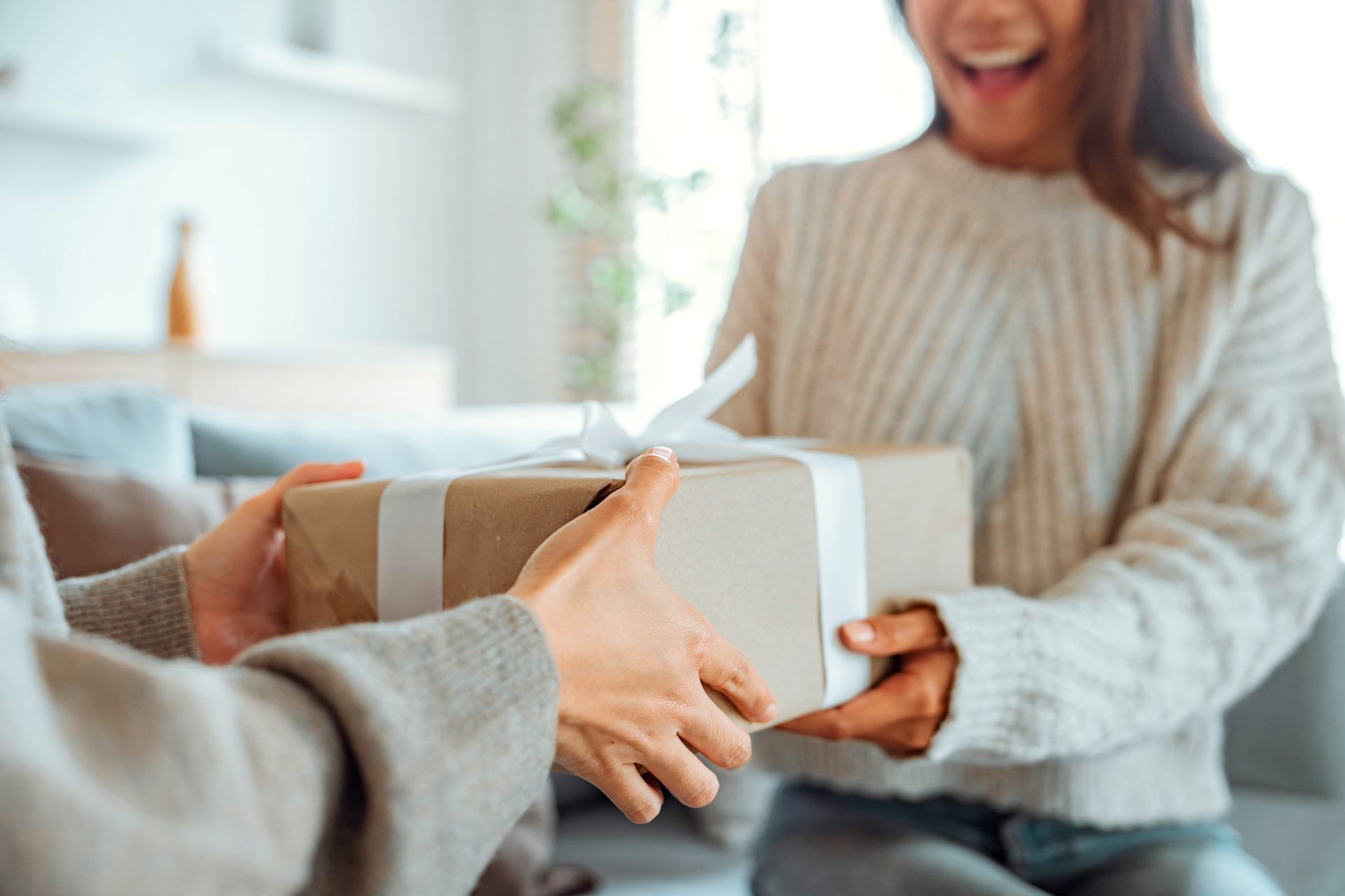 Hands exchanging a wrapped gift in a bright living room, with a smiling person in the background
