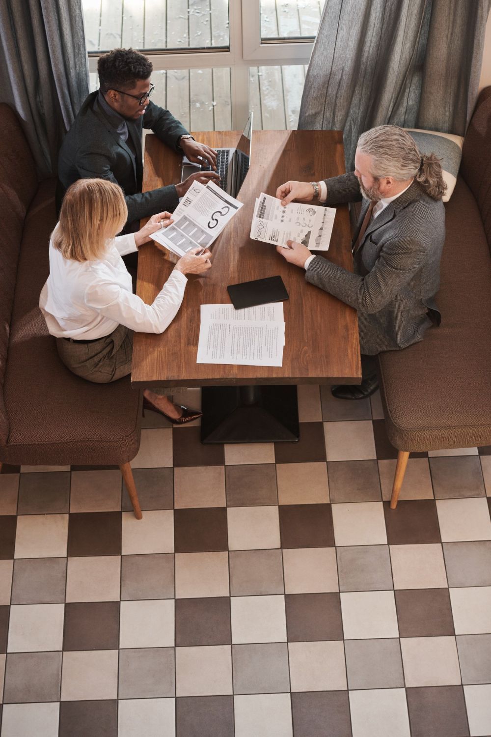 Three people sit at a wooden table in a booth with a checkered floor, reviewing documents and working on a laptop.