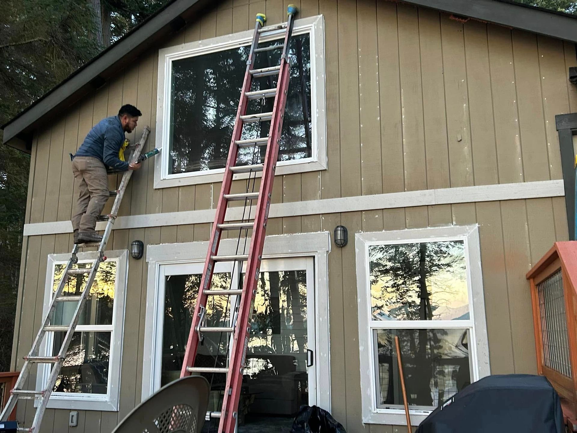 A man on a ladder paints the exterior of a tan house. Two windows are visible.
