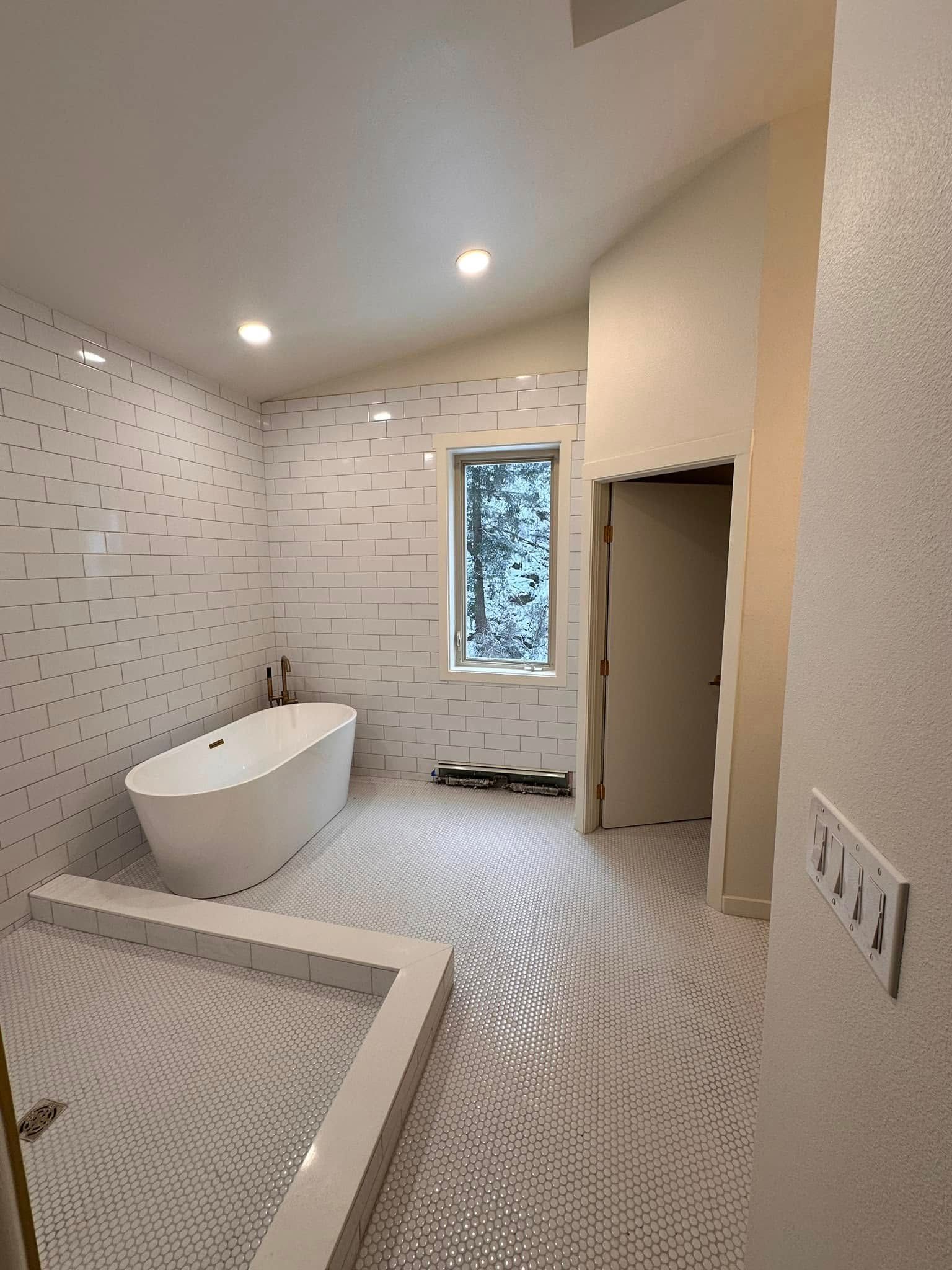 Bathroom with white tile walls and floor, a freestanding tub, and a doorway.