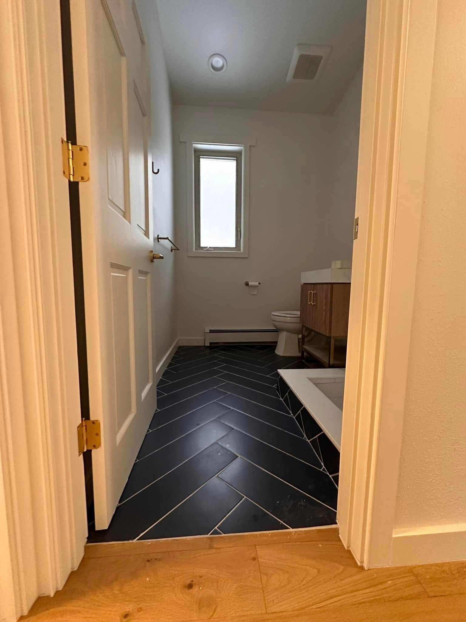 Bathroom with black herringbone floor, white walls, open door, and wooden vanity.