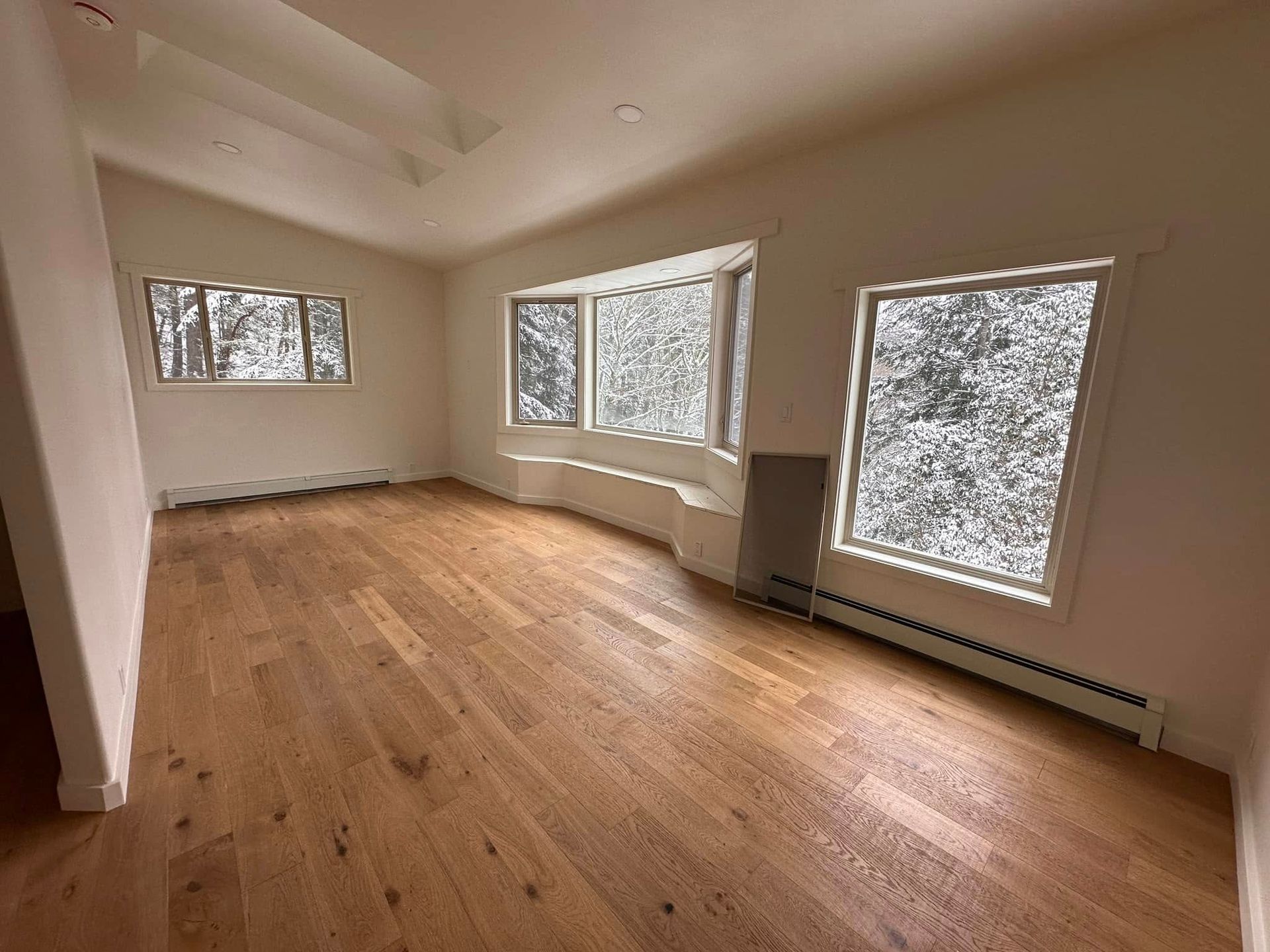 Empty room with wood floor, white walls, and large windows with a forest view.