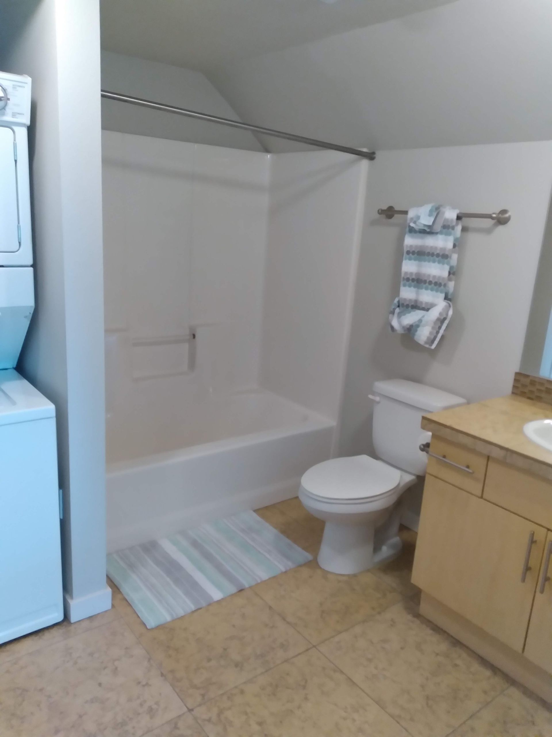 Bathroom with white tub-shower, toilet, wood vanity, and striped towels on beige tile floor.