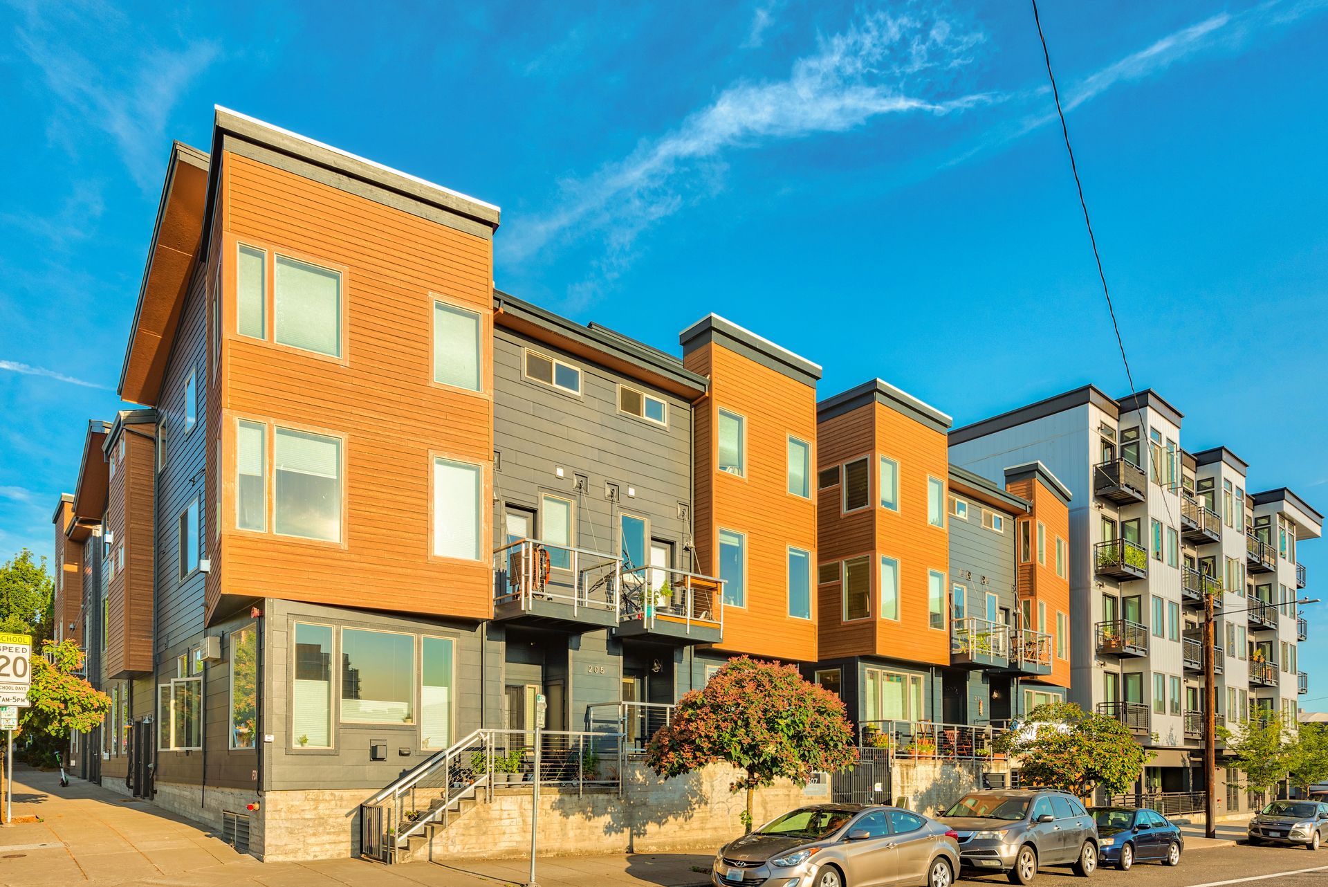 Modern apartment buildings with orange and gray facades along a sunny street, with parked cars and blue sky