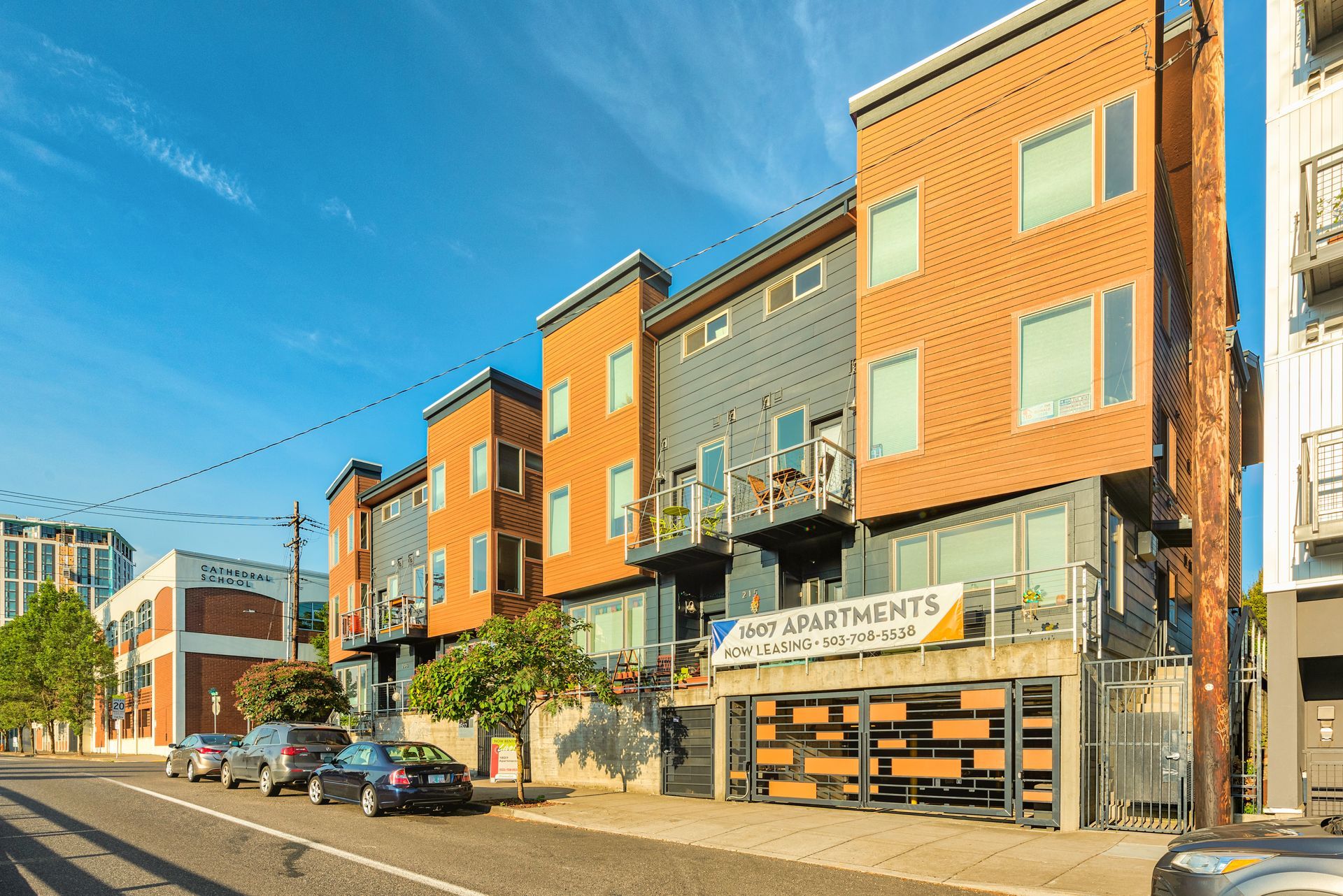 Modern apartment building with orange and gray facade, balconies, and parked cars on a sunny street
