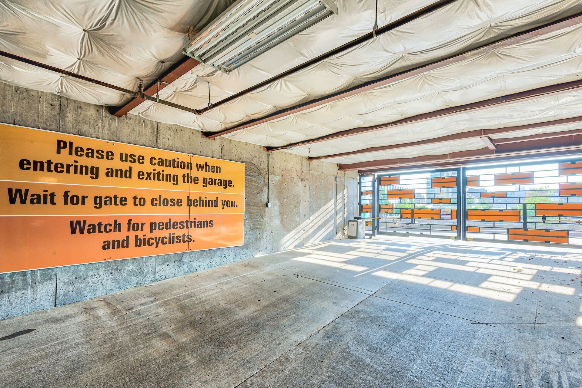 Parking garage entrance with a yellow safety sign, concrete ceiling, and sunlight through the open gate