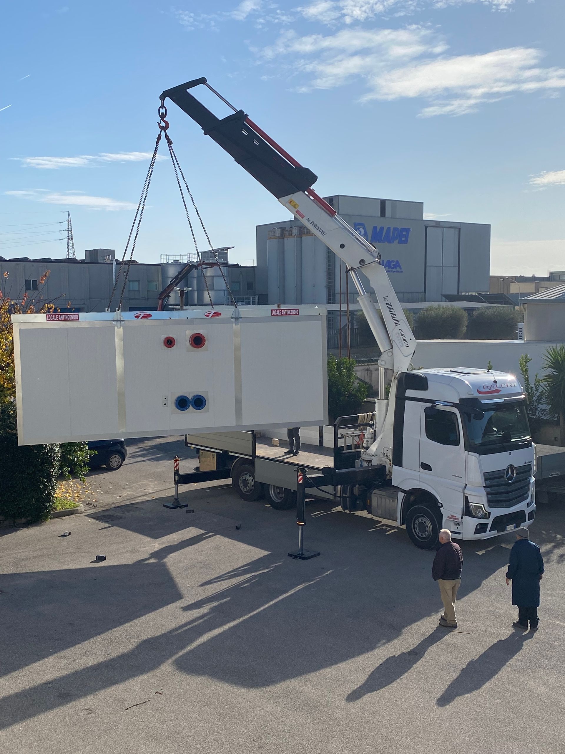 Un carro attrezzi solleva un camion blu accanto a una gru gialla in un'area industriale.