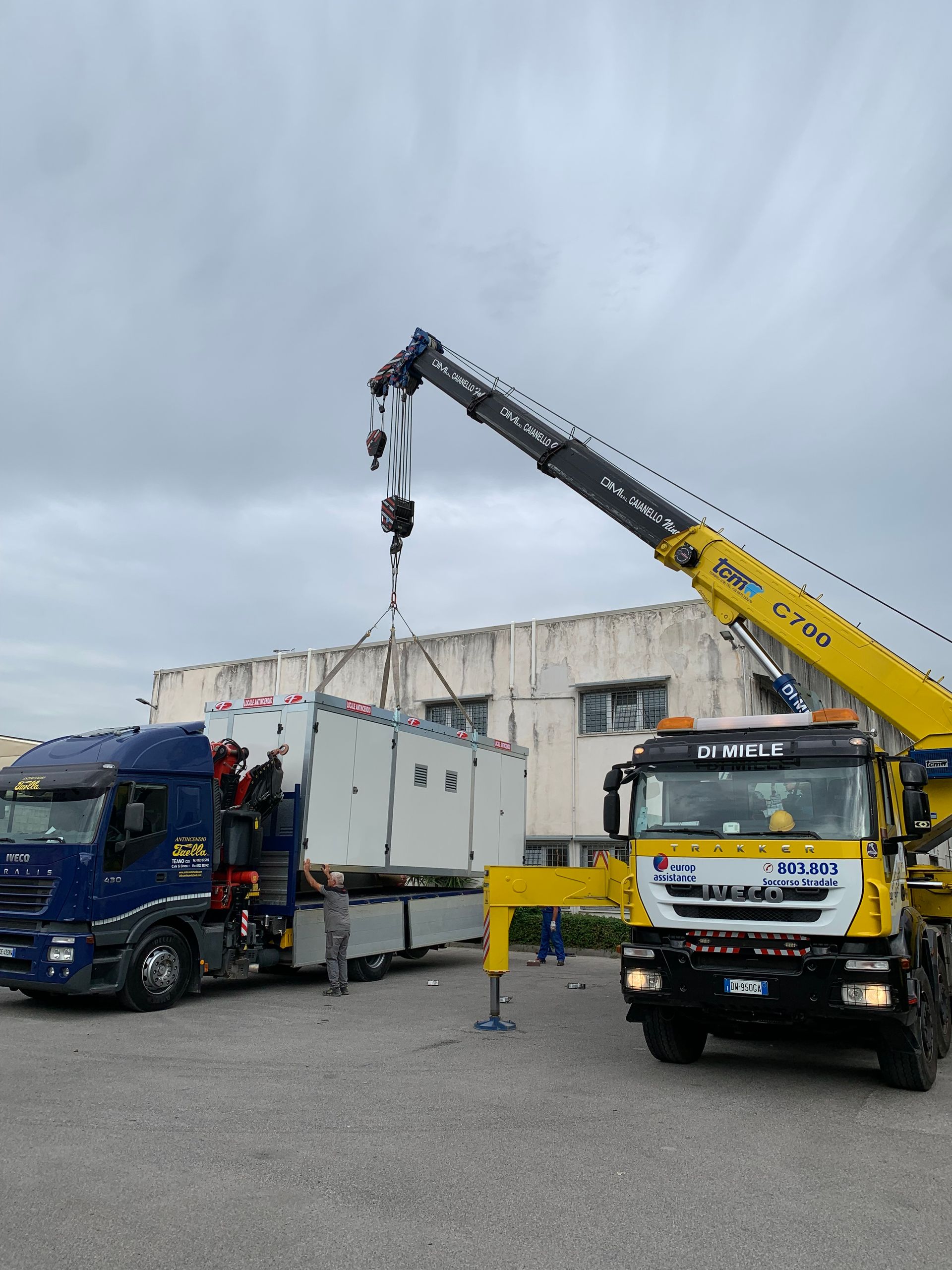 Un carro attrezzi solleva un camion blu accanto a una gru gialla in un'area industriale.
