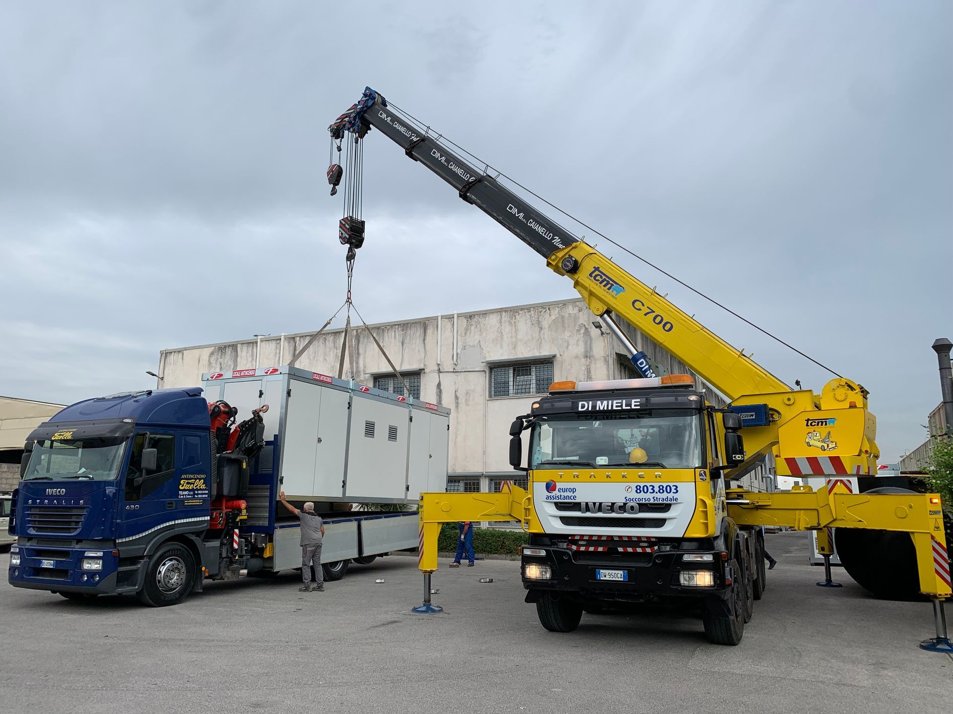 Un carro attrezzi solleva un camion blu accanto a una gru gialla in un'area industriale.