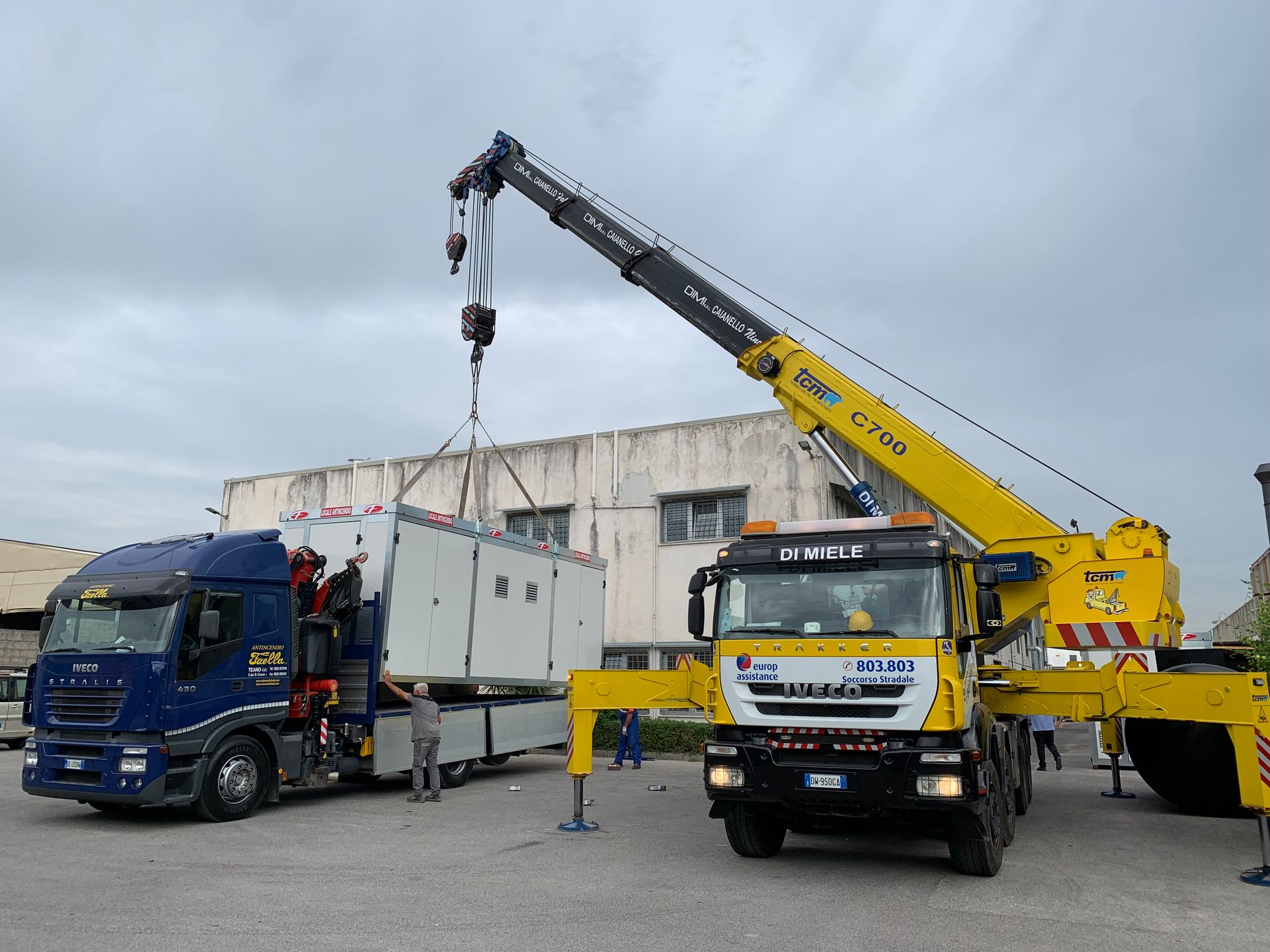 Un carro attrezzi solleva un camion blu accanto a una gru gialla in un'area industriale.