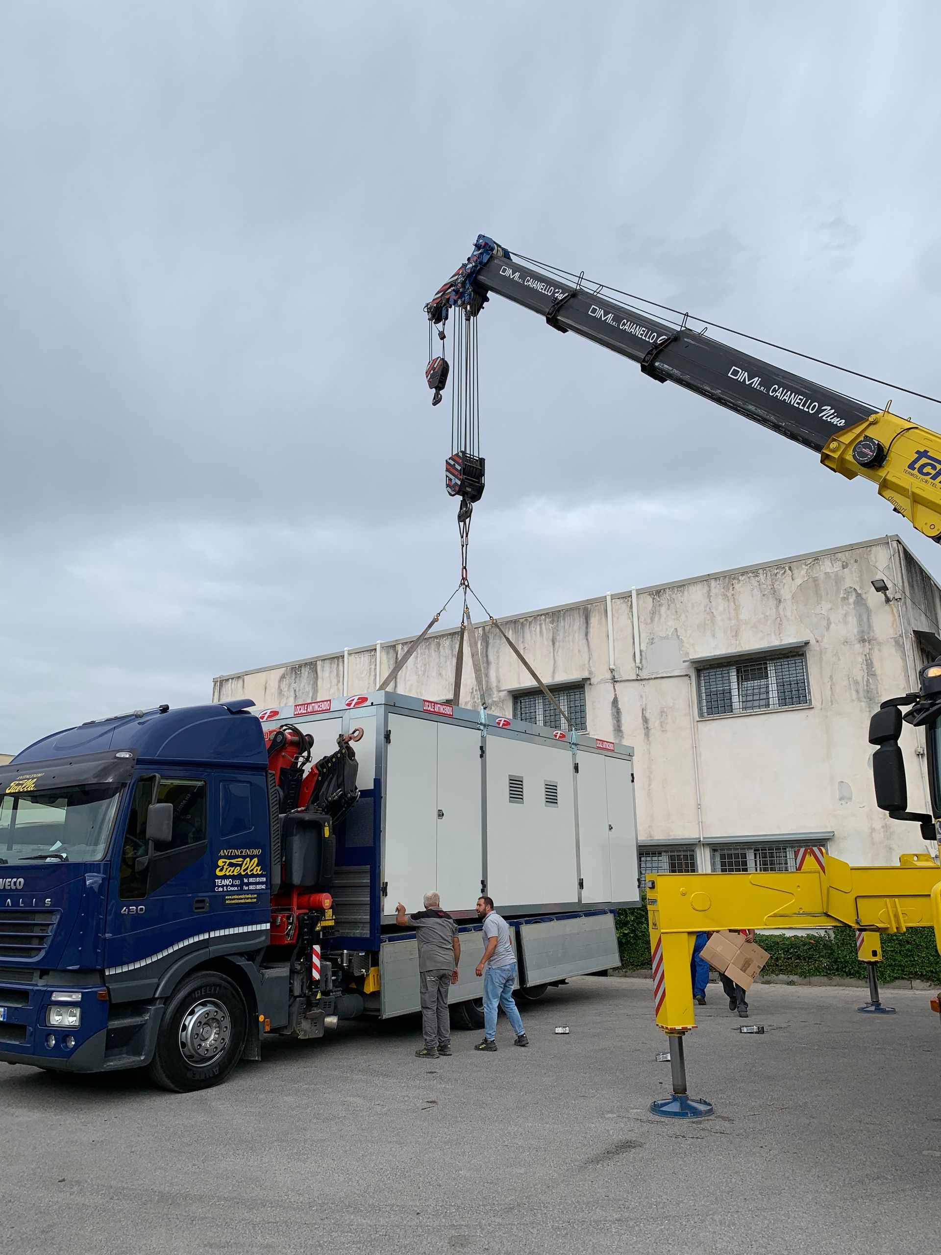 Un carro attrezzi solleva un camion blu accanto a una gru gialla in un'area industriale.
