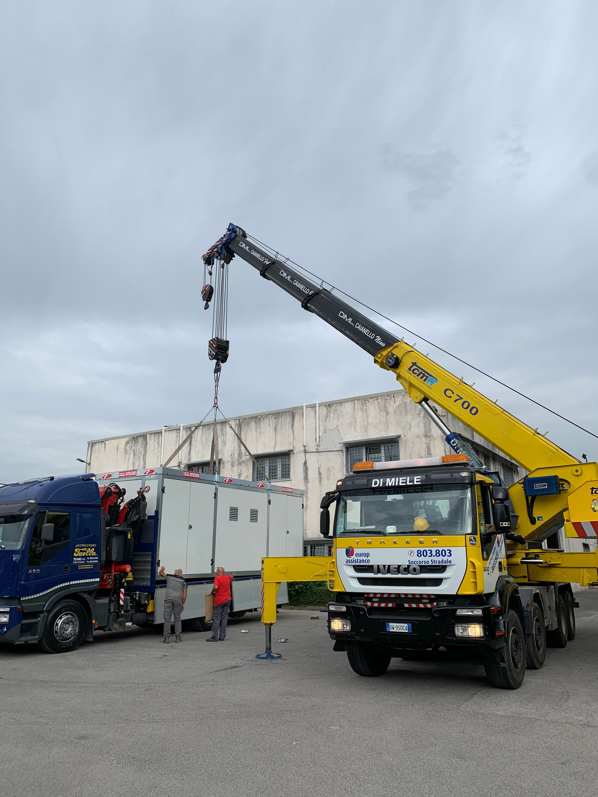 Un carro attrezzi solleva un camion blu accanto a una gru gialla in un'area industriale.