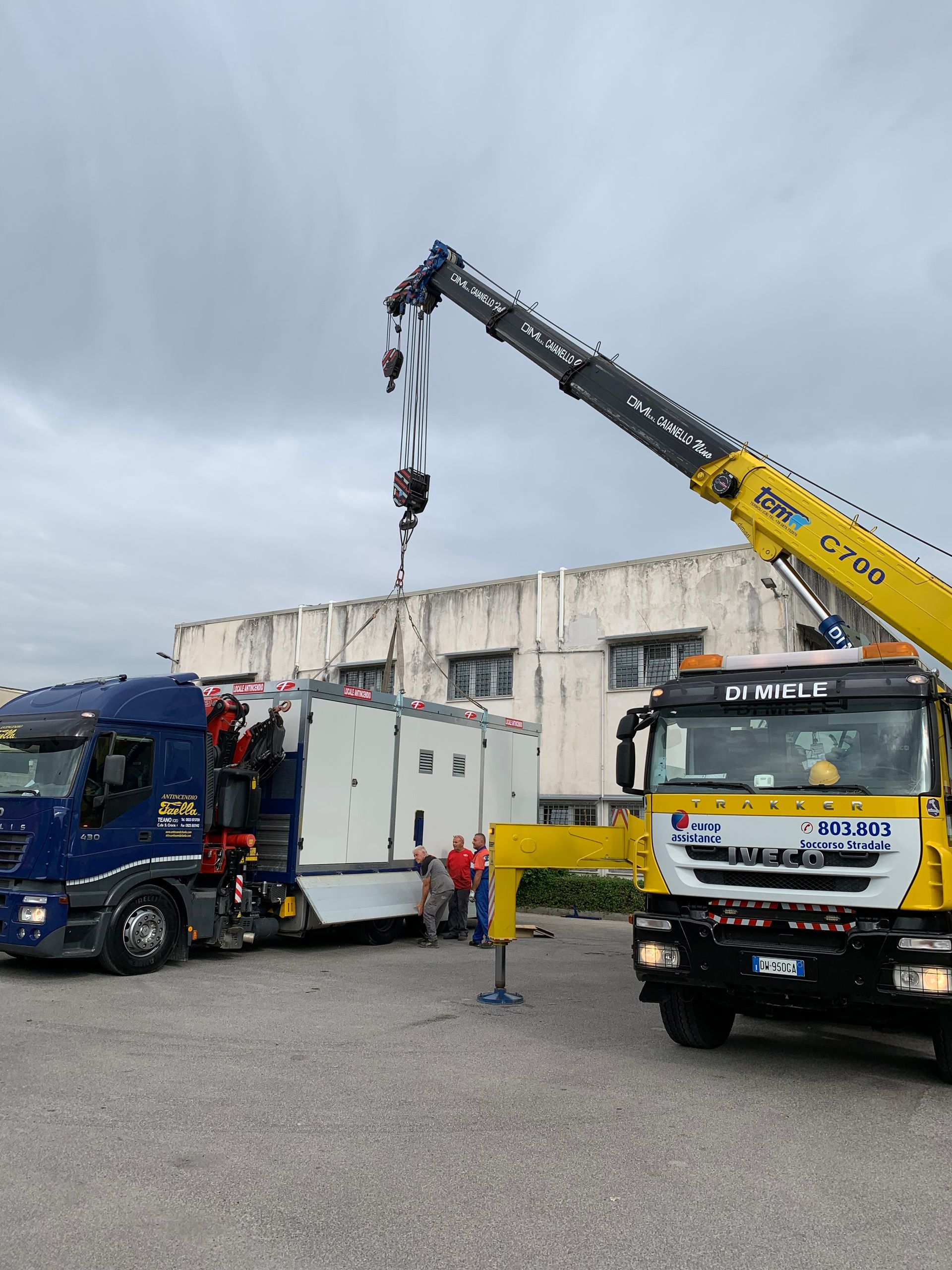Un carro attrezzi solleva un camion blu accanto a una gru gialla in un'area industriale.