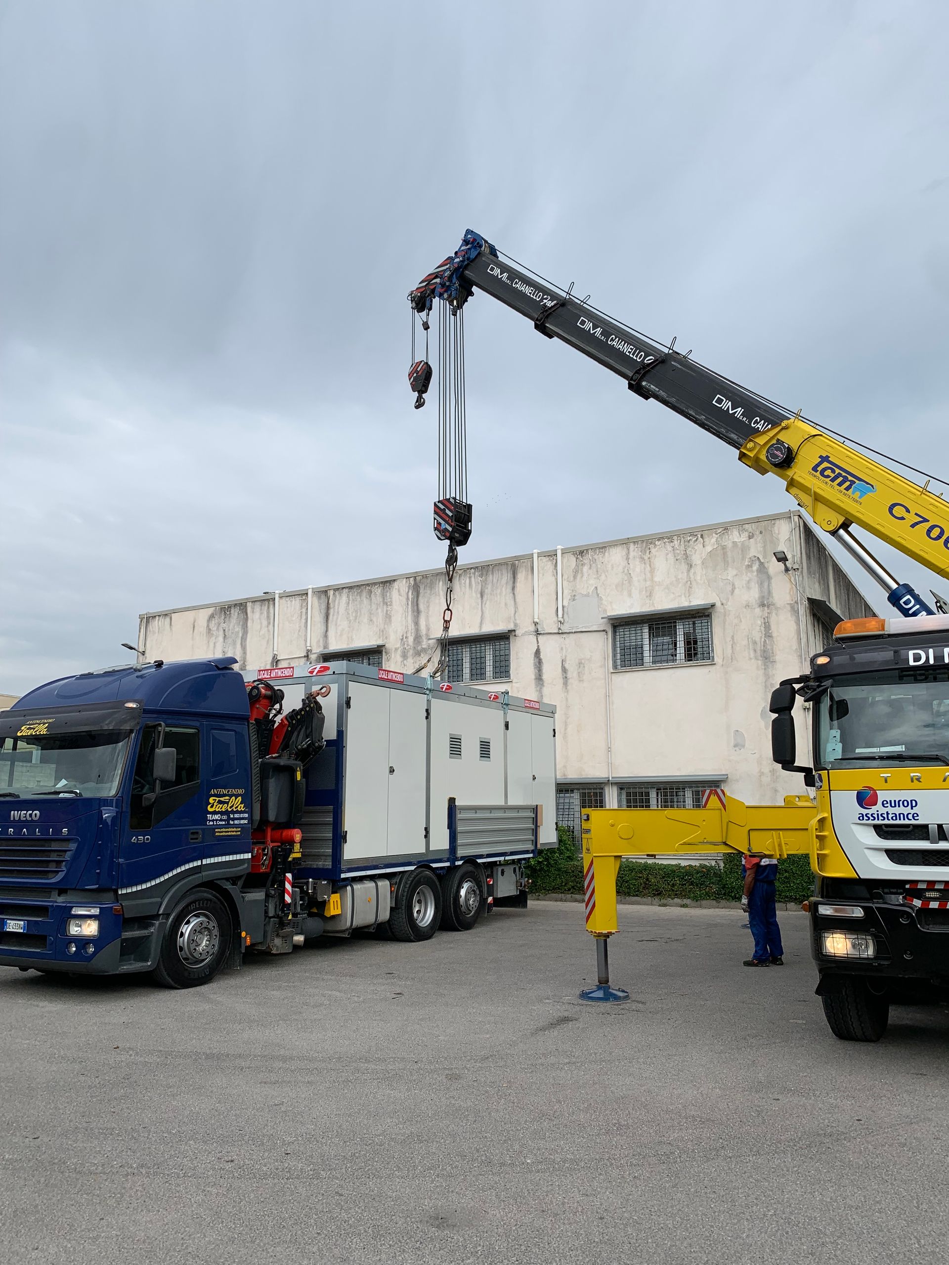 Un carro attrezzi solleva un camion blu accanto a una gru gialla in un'area industriale.