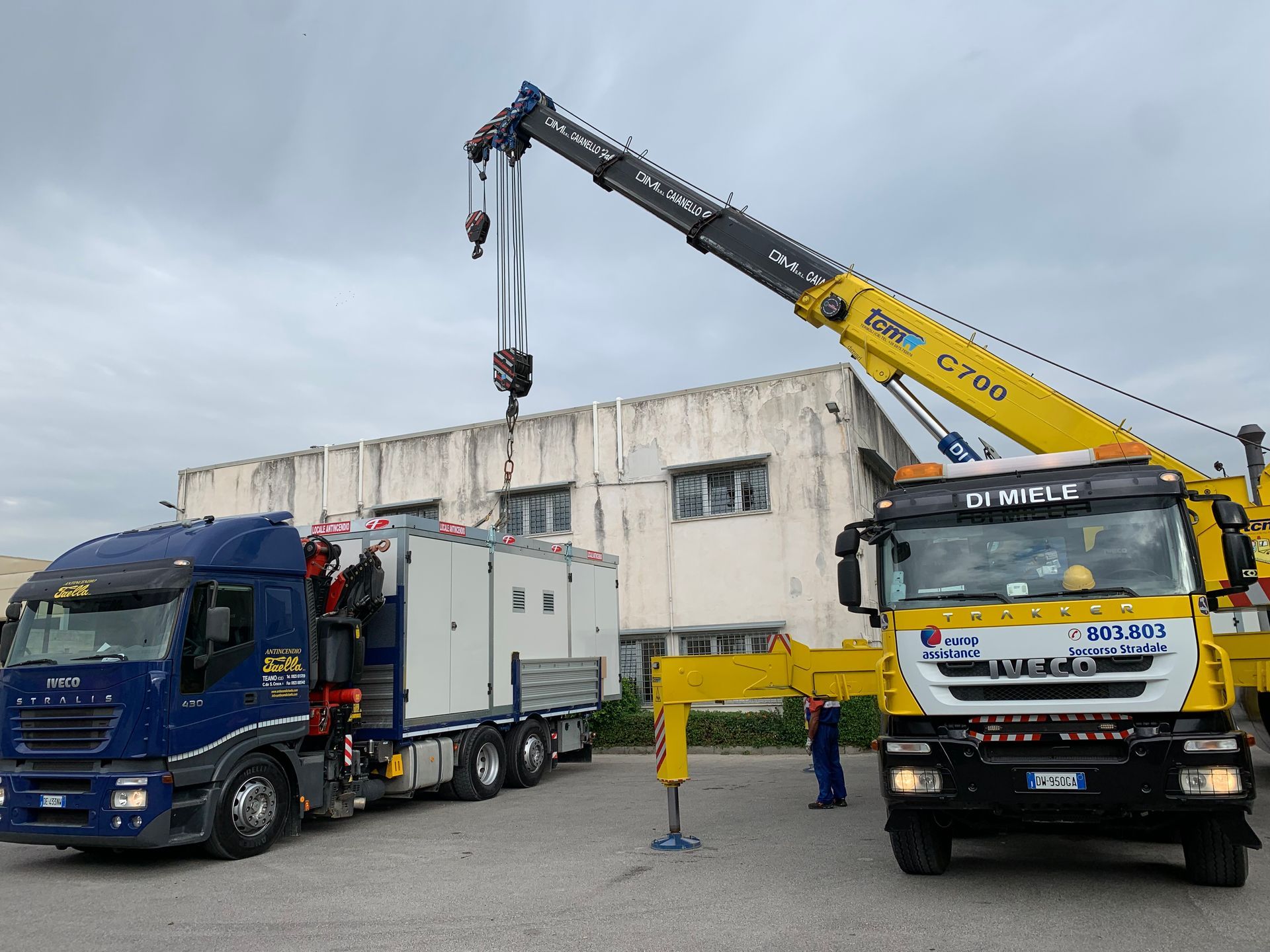 Un carro attrezzi solleva un camion blu accanto a una gru gialla in un'area industriale.