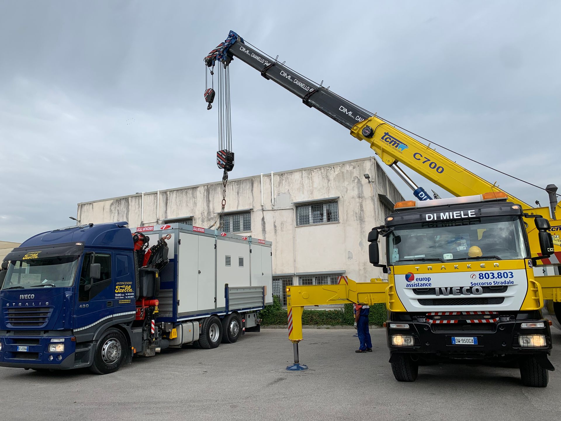Un carro attrezzi solleva un camion blu accanto a una gru gialla in un'area industriale.