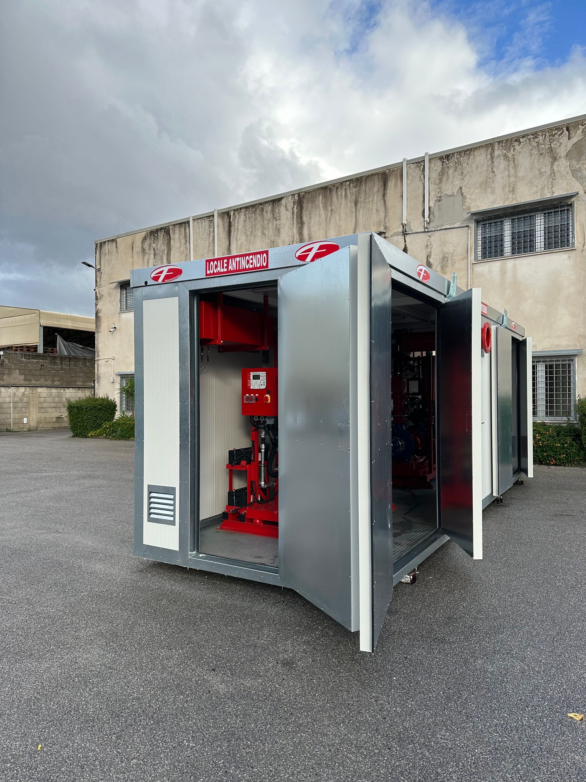 Armadietto di emergenza rosso in un rifugio metallico aperto all'esterno di un edificio in cemento sotto un cielo nuvoloso.