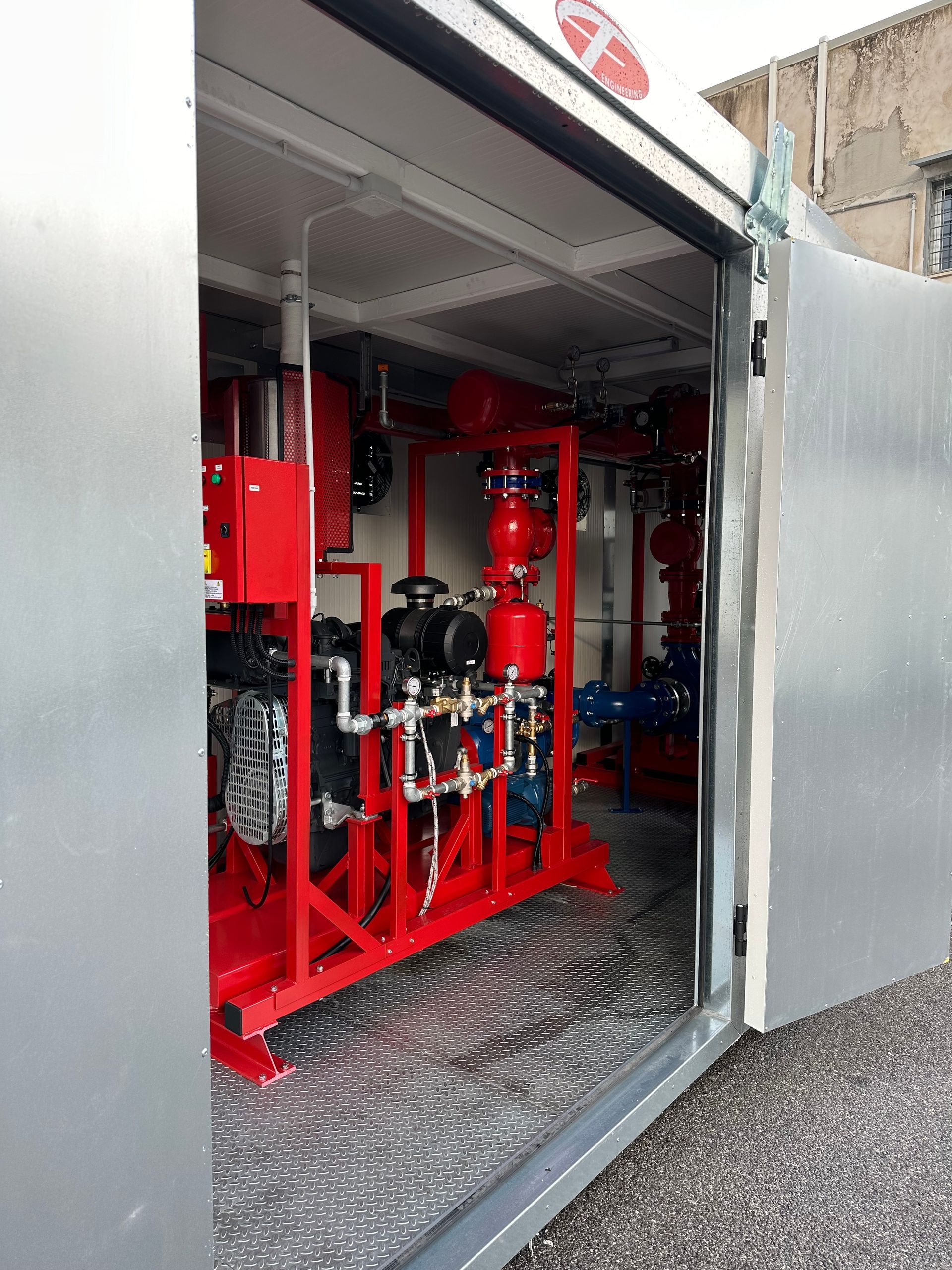 Tubature e macchinari industriali rossi all'interno di un container metallico su un pavimento in lamiera mandorlata.