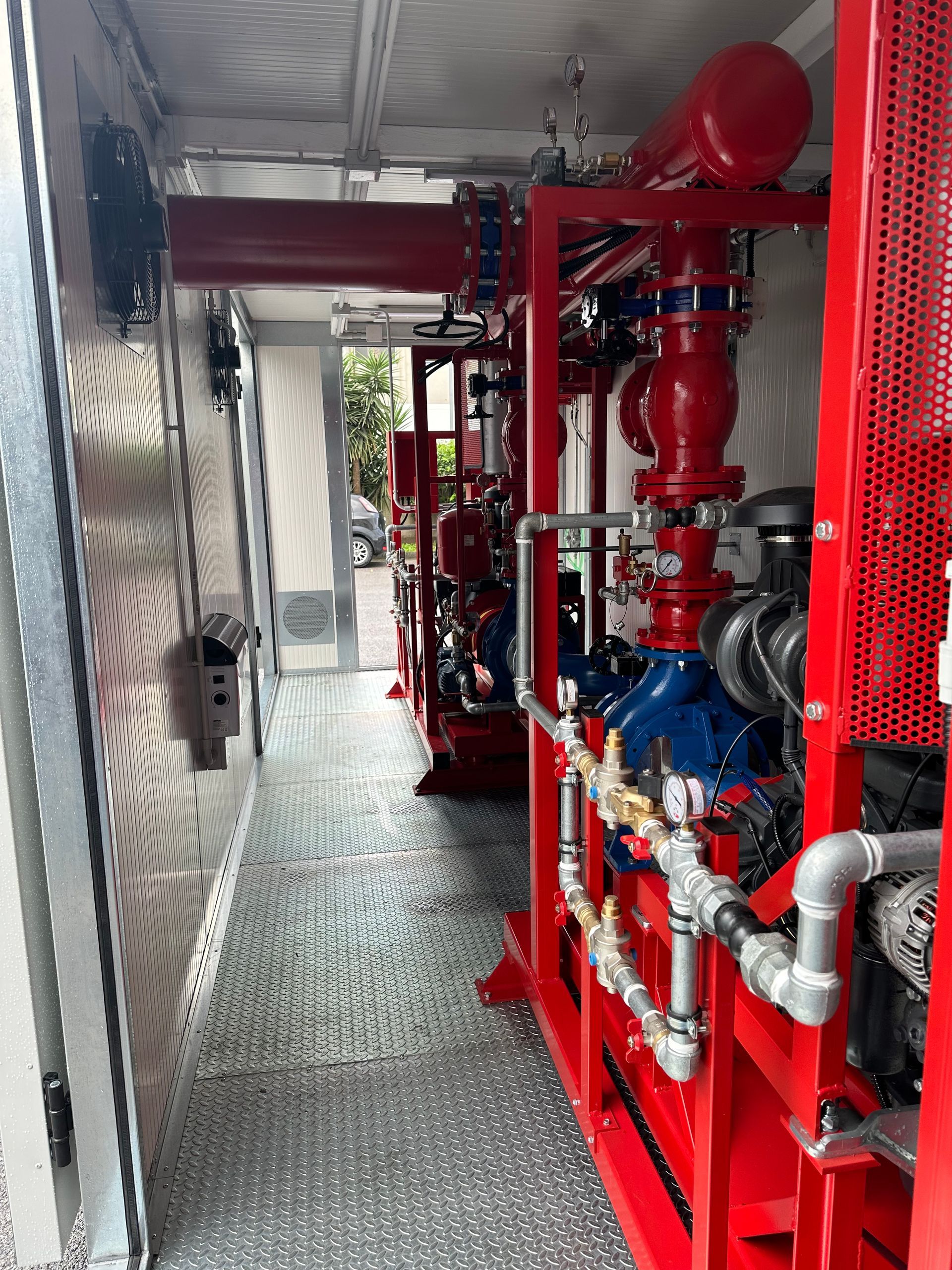 Tubature e valvole antincendio rosse in uno stretto corridoio industriale con attrezzature e ringhiera a rete.
