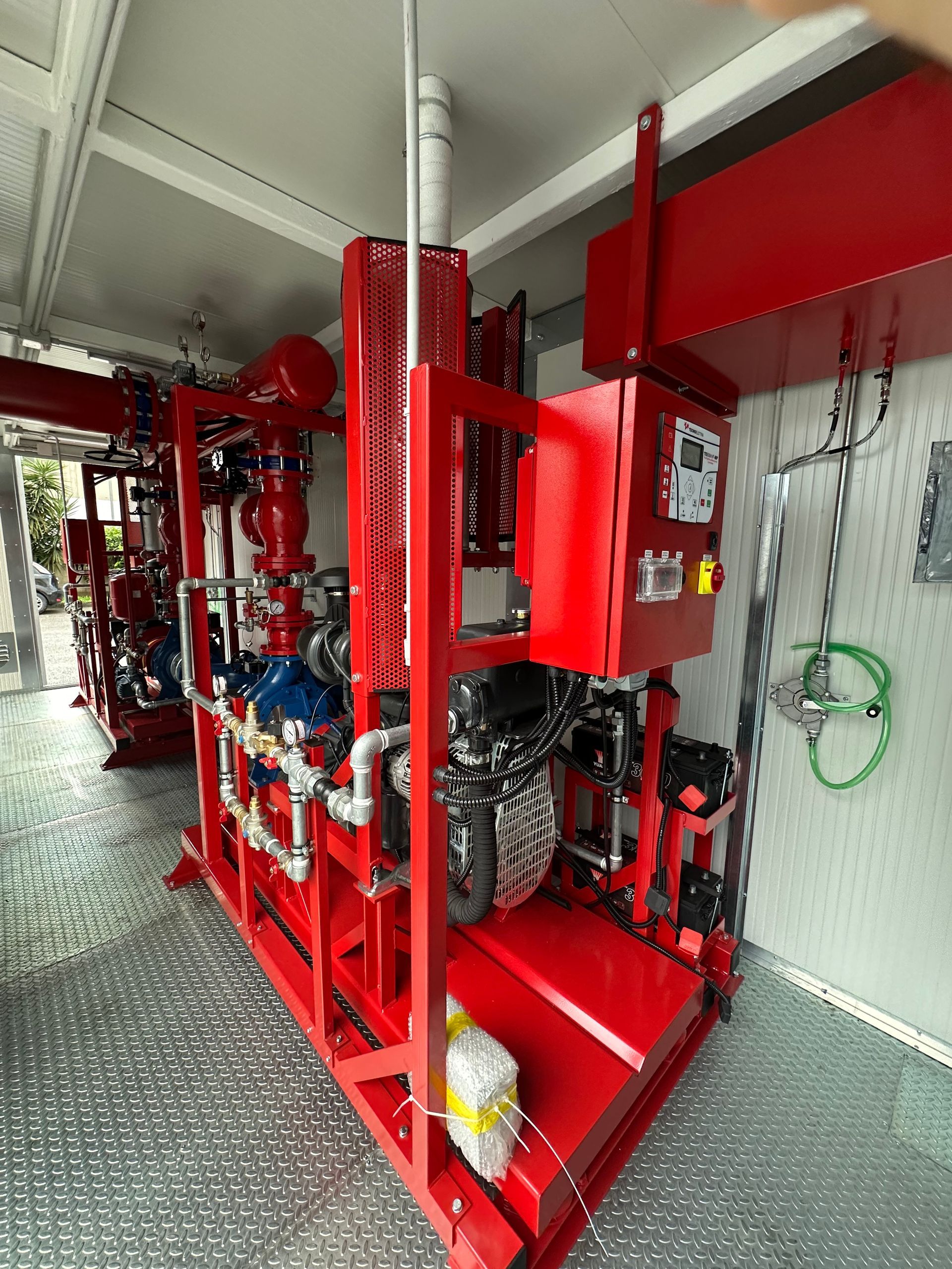 Tubature e valvole antincendio rosse in uno stretto corridoio industriale con attrezzature e ringhiera a rete.