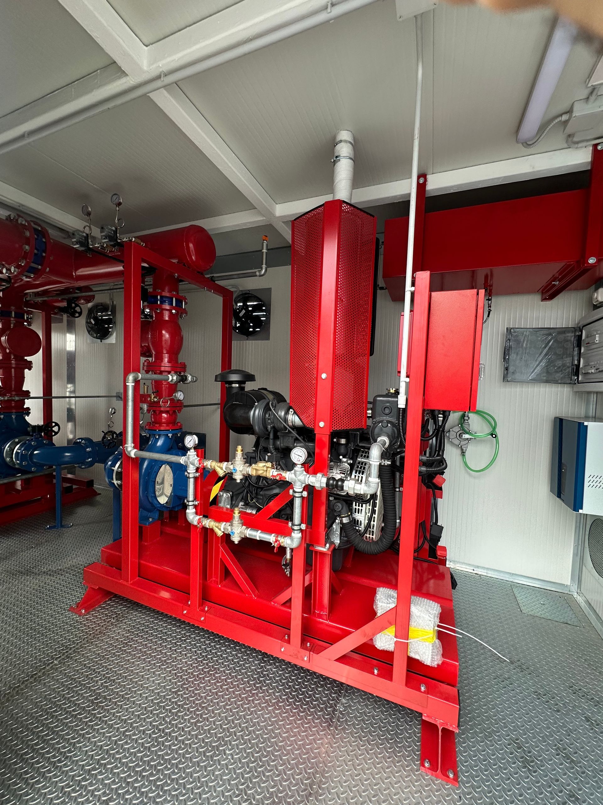 Tubature e valvole antincendio rosse in uno stretto corridoio industriale con attrezzature e ringhiera a rete.