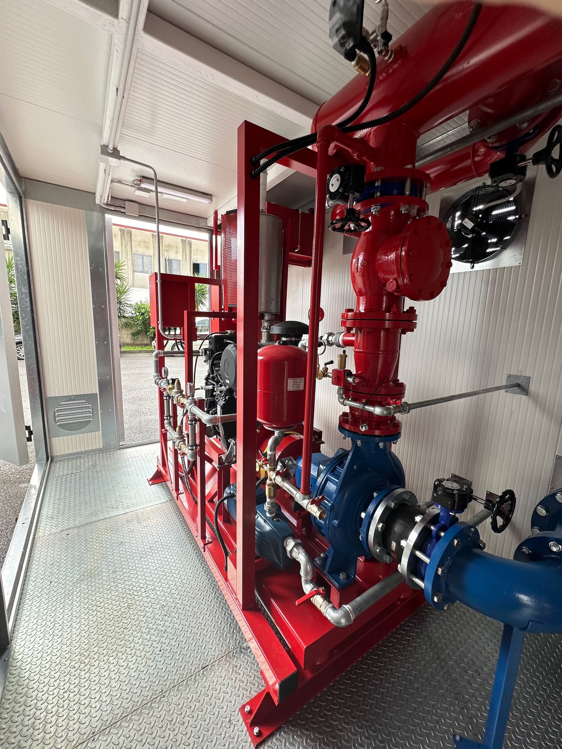 Tubature e valvole antincendio rosse in uno stretto corridoio industriale con attrezzature e ringhiera a rete.