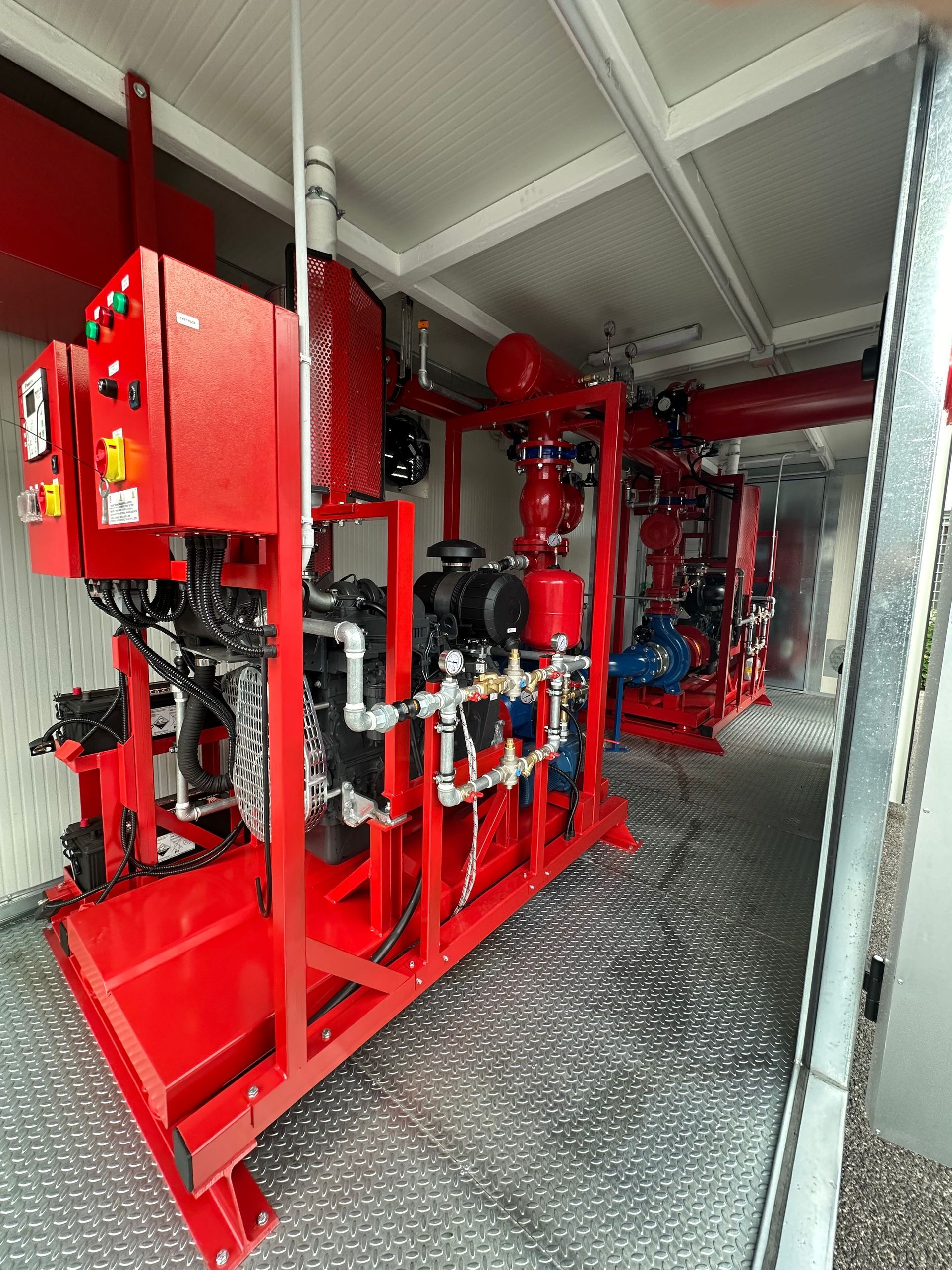 Tubature e valvole antincendio rosse in uno stretto corridoio industriale con attrezzature e ringhiera a rete.