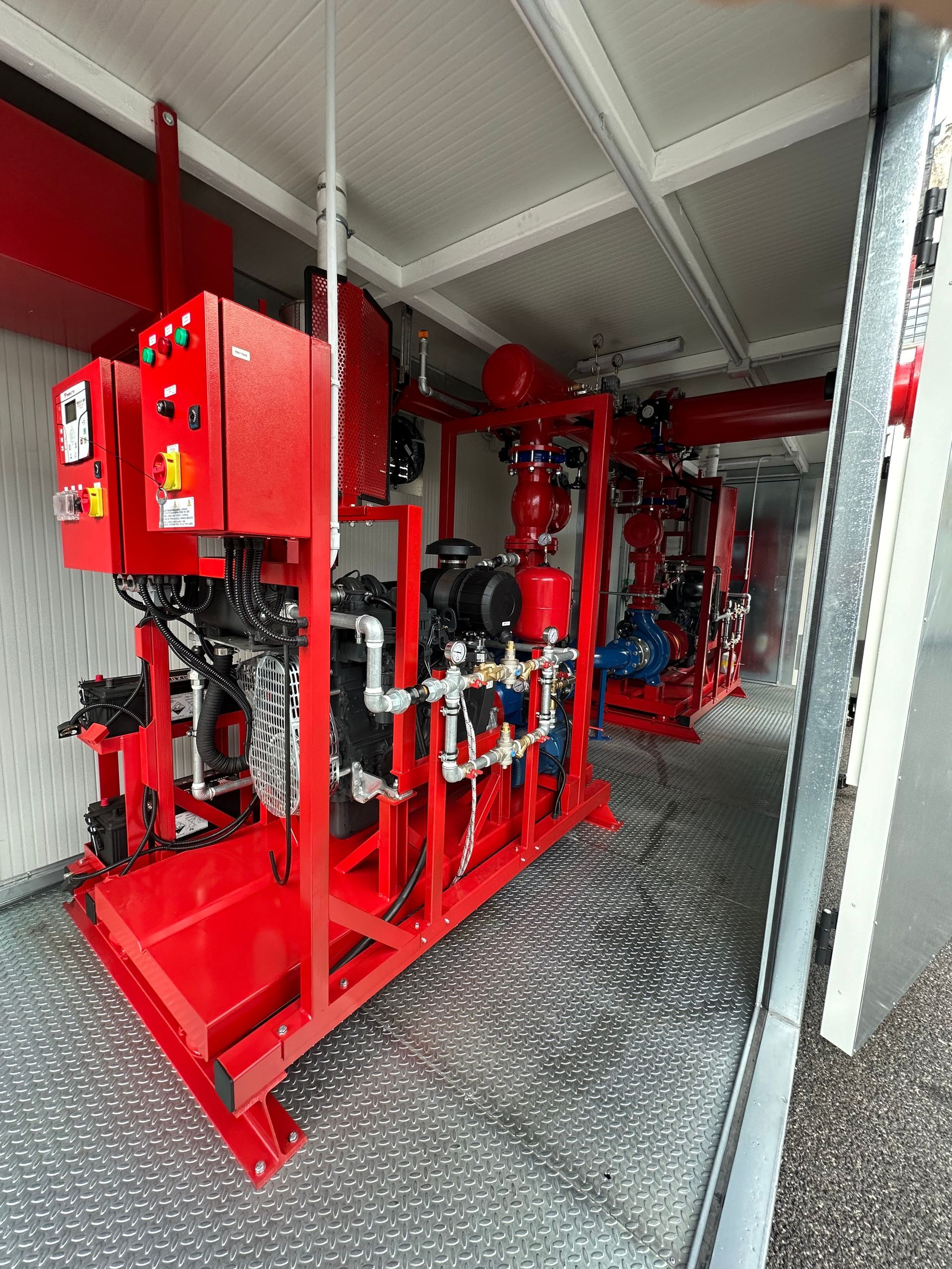 Tubature e valvole antincendio rosse in uno stretto corridoio industriale con attrezzature e ringhiera a rete.