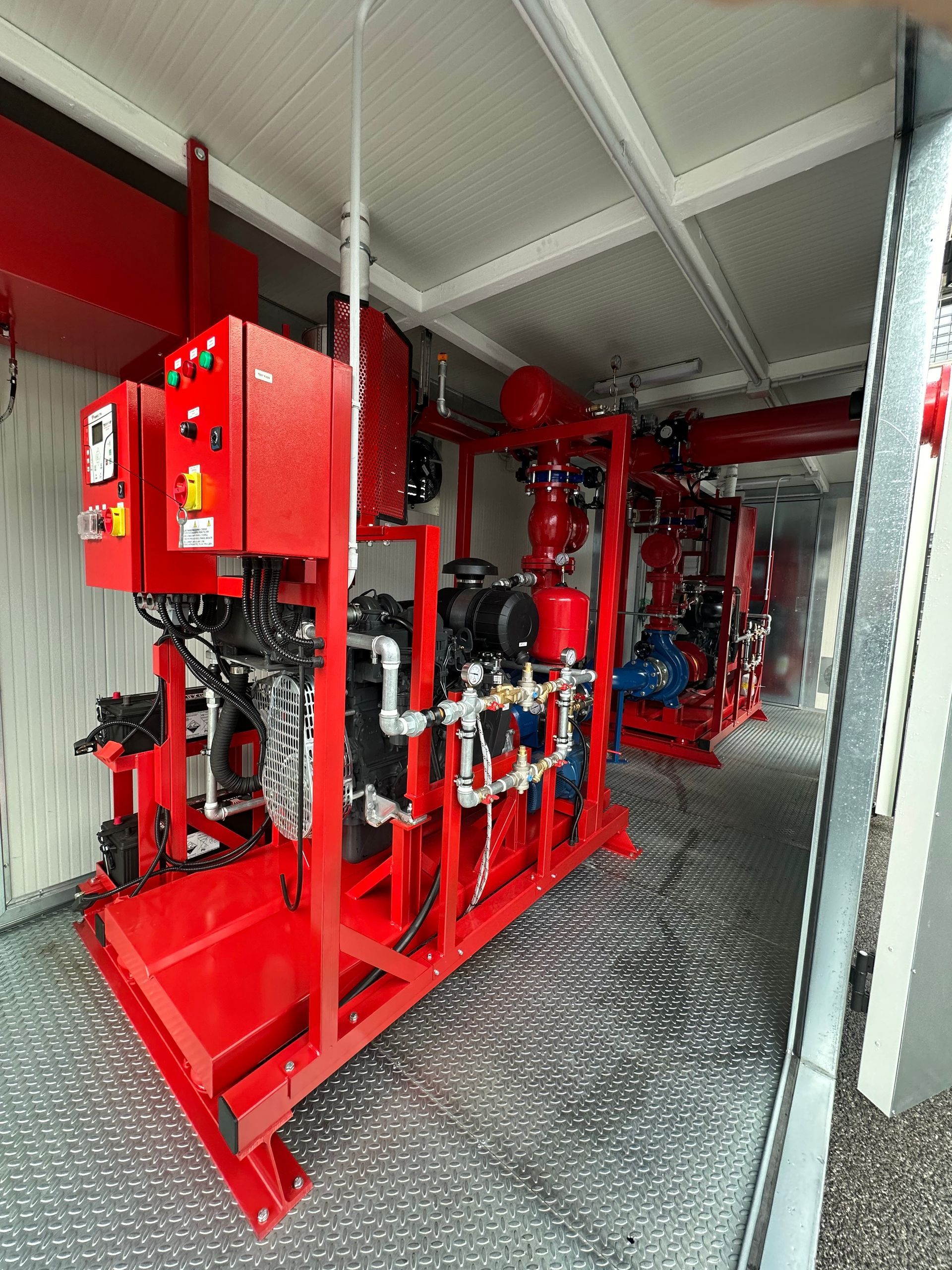 Tubature e valvole antincendio rosse in uno stretto corridoio industriale con attrezzature e ringhiera a rete.