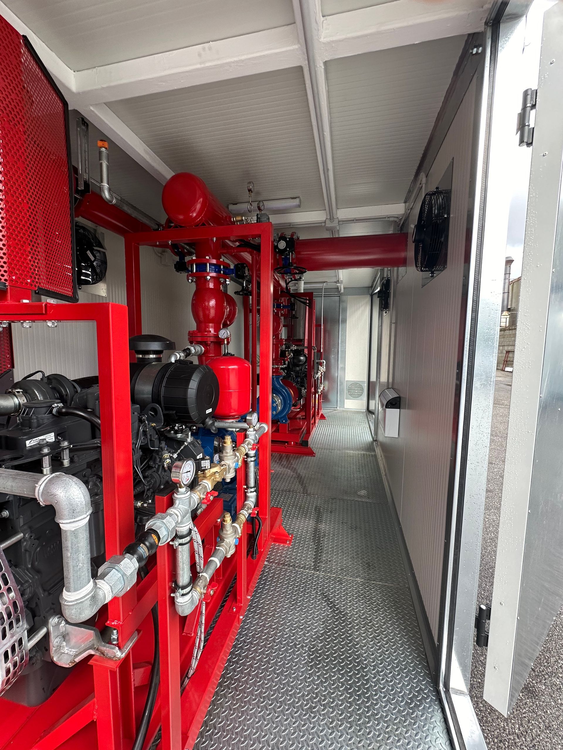 Tubature e valvole antincendio rosse in uno stretto corridoio industriale con attrezzature e ringhiera a rete.