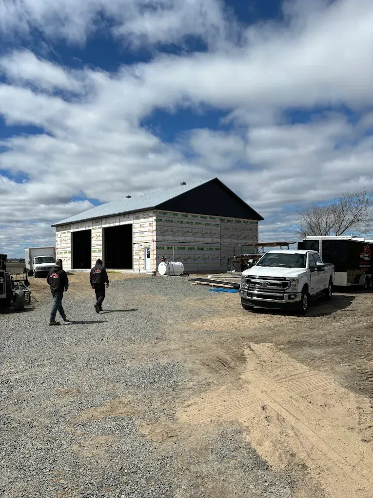 Chantier de construction avec une maison partiellement construite, deux ouvriers et une camionnette blanche sous un ciel nuageux