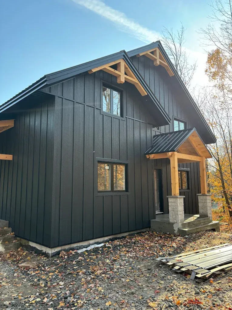 Cabane moderne noire avec boiseries et petite véranda dans une zone boisée, avec du gravier et du bois empilé à proximité.