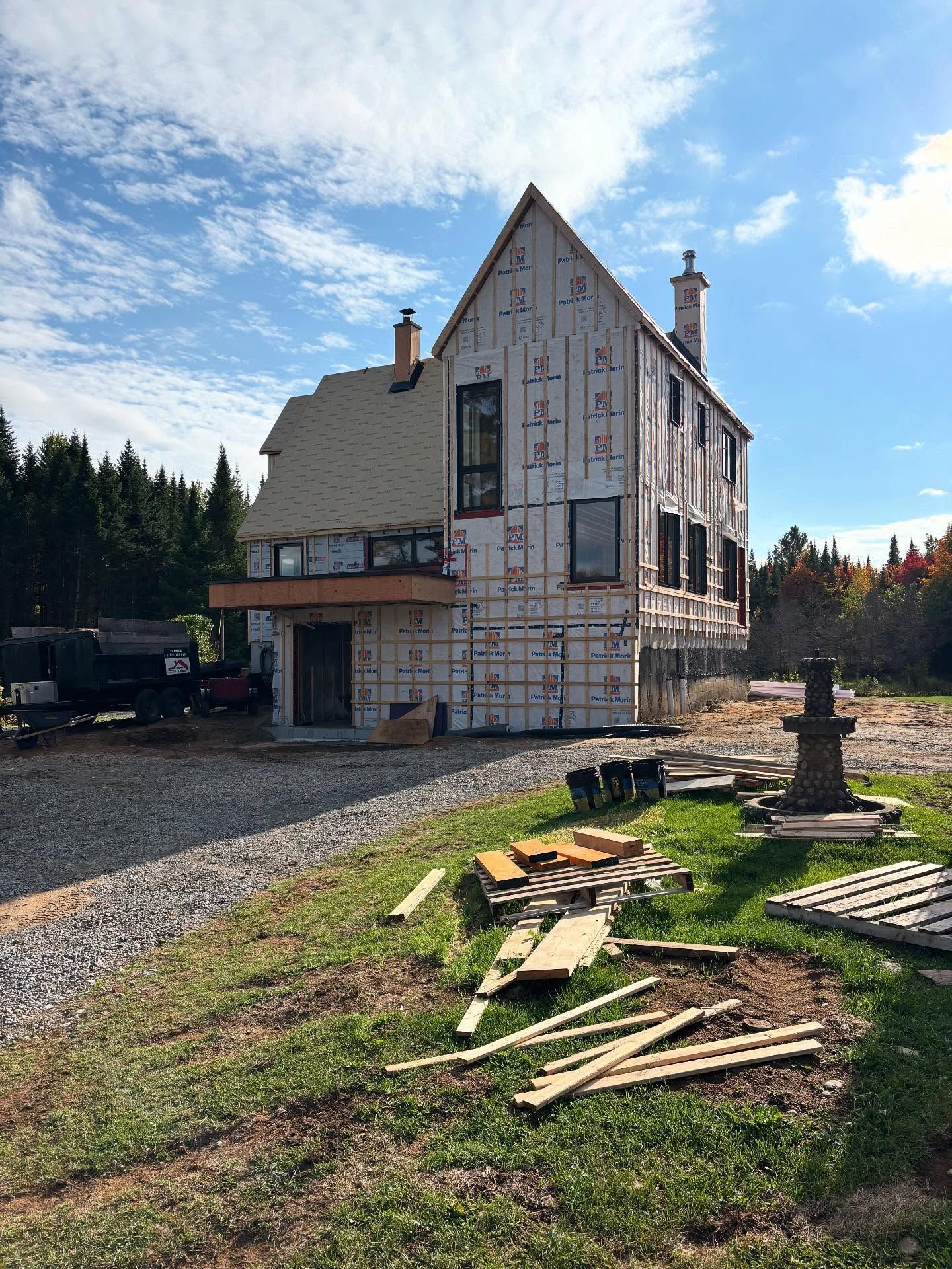 Maison partiellement construite avec bardage apparent, allée de gravier et bois d'œuvre éparpillés sous un ciel bleu